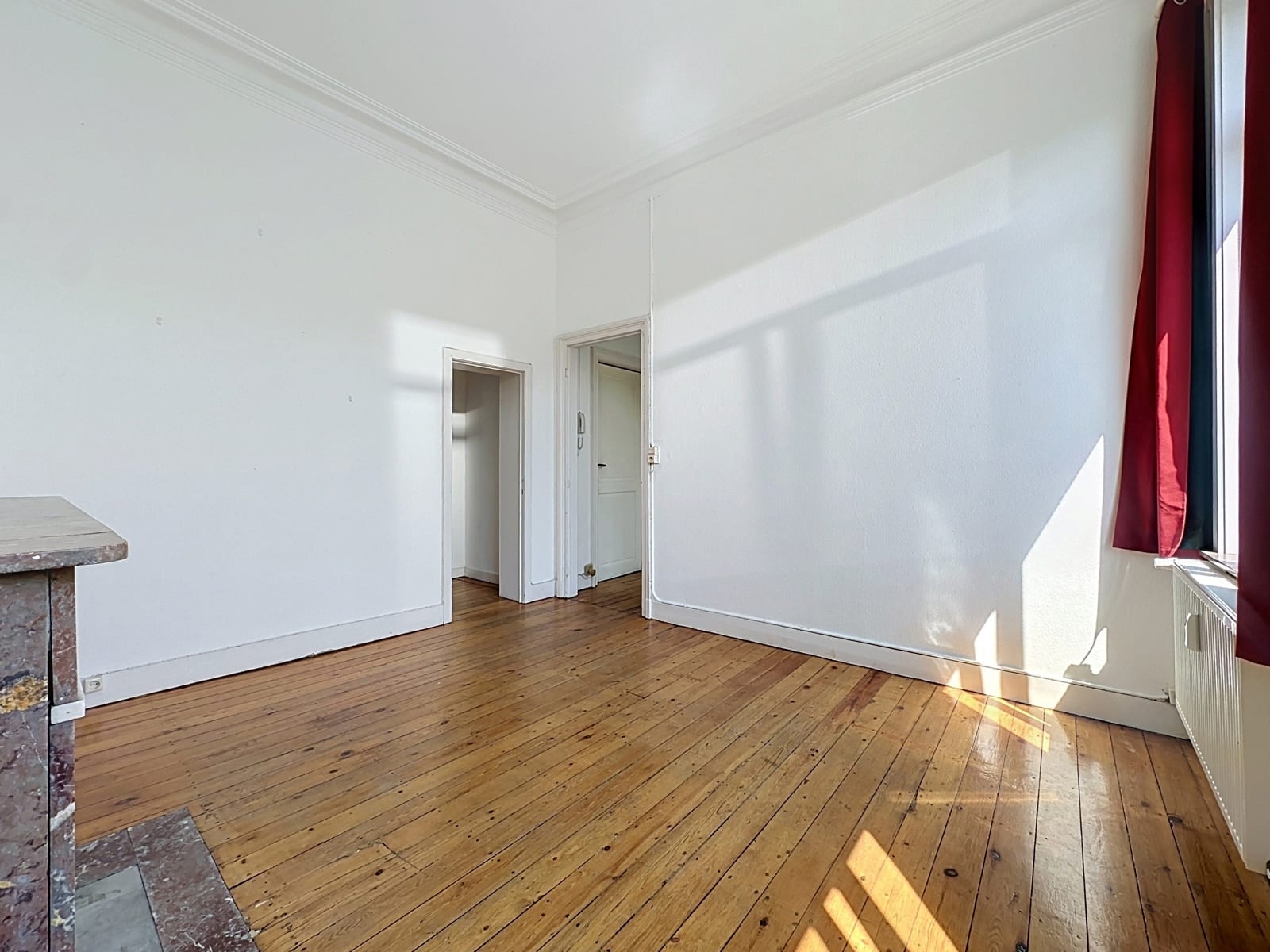 Salon Lumineux avec Parquet Ancien – You Real Estate Agency Salon lumineux d’appartement avec parquet ancien, moulures blanches et rideaux rouges.