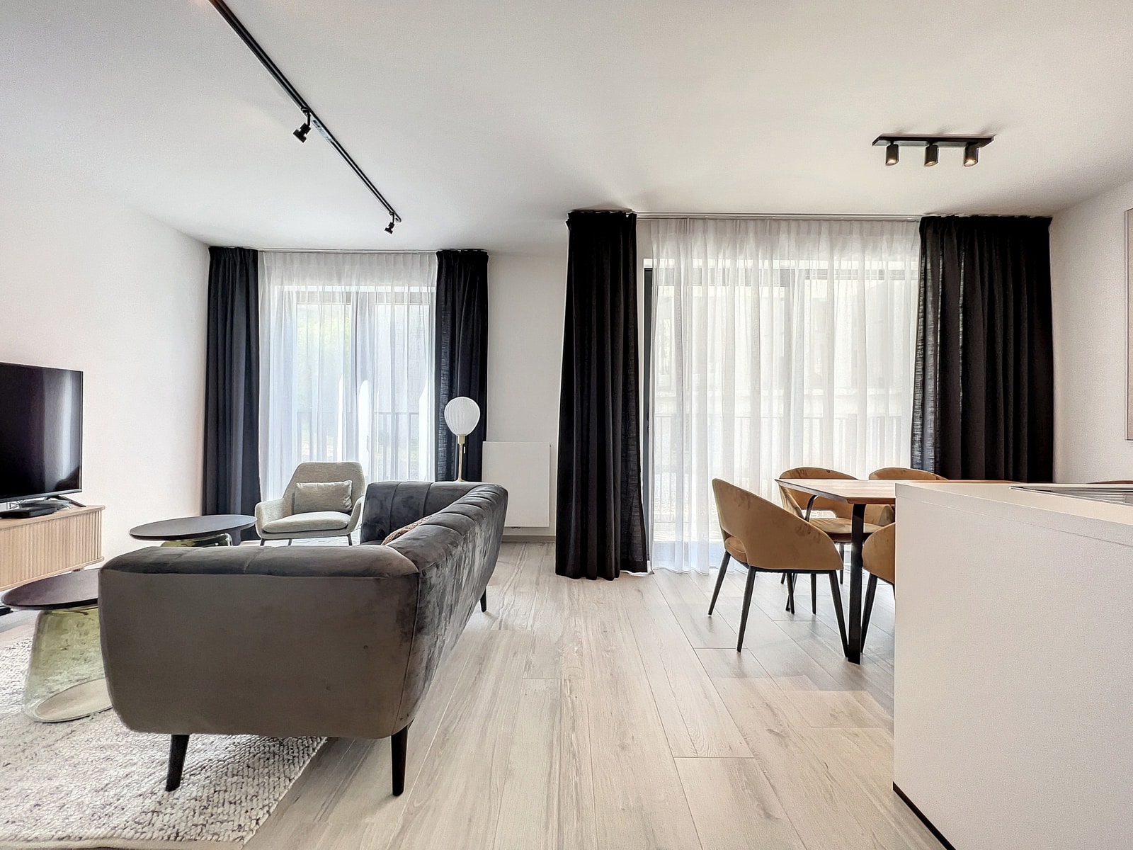 whise photo 2 Bright living room with a gray sectional sofa, round coffee tables, and a TV on a low stand beside large windows with sheer white and black curtains.