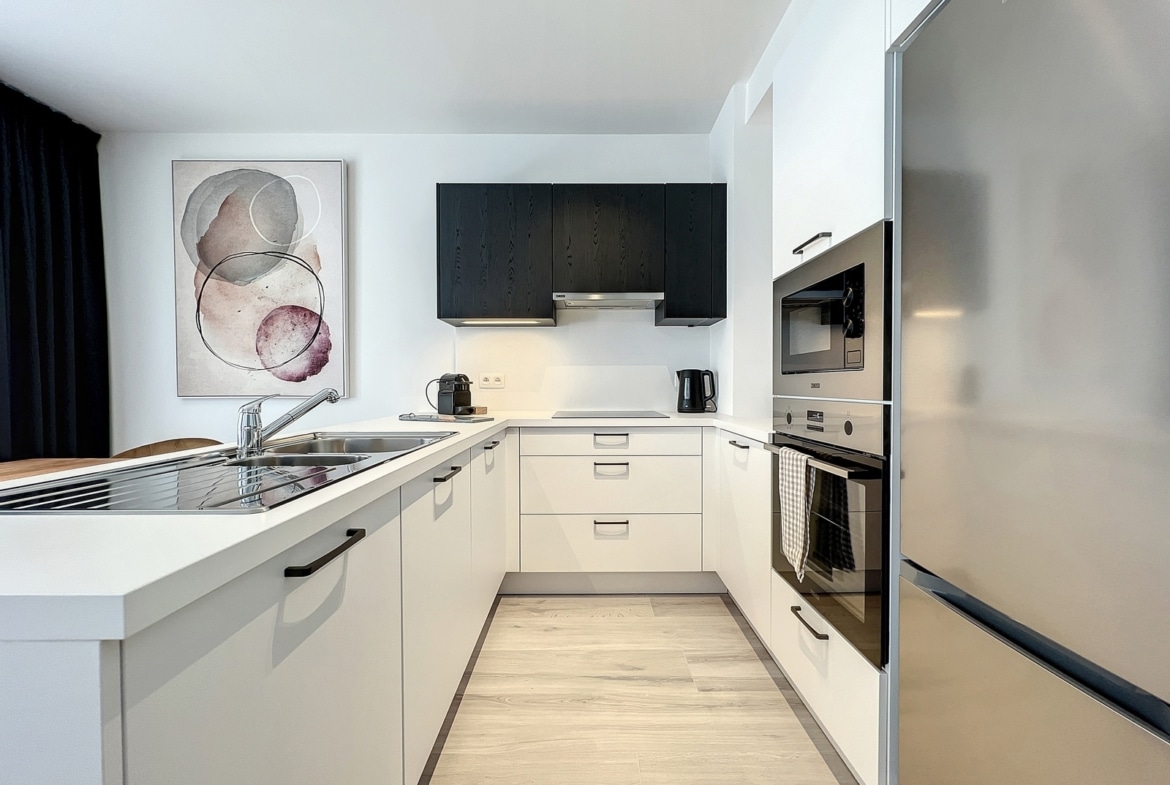 Modern white kitchen with an island sink, black wall cabinets, and stainless steel appliances beneath a row of white cabinets. An abstract art print hangs on the left wall.