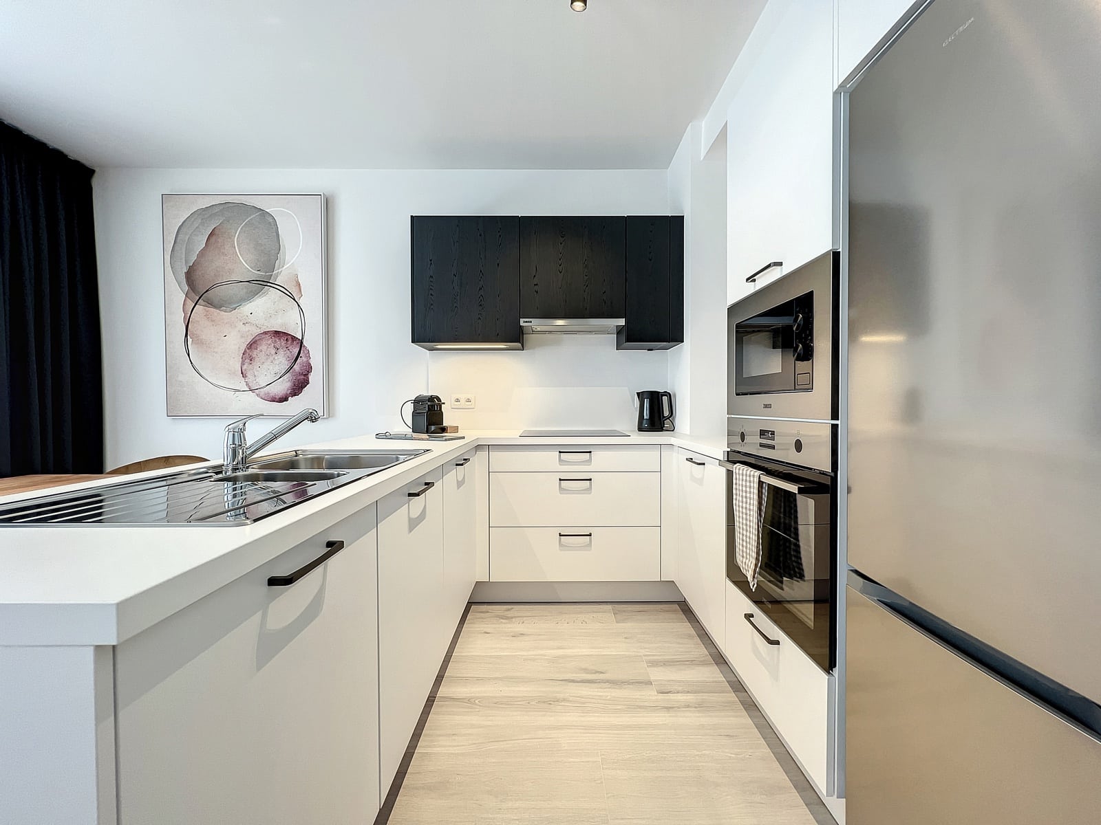 whise photo 3 Modern white kitchen with an island sink, black wall cabinets, and stainless steel appliances beneath a row of white cabinets. An abstract art print hangs on the left wall.