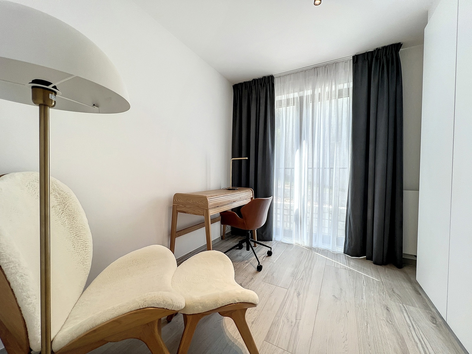 whise photo 8 Minimalist home office nook with a wooden desk, brass lamp, and a reddish-brown swivel chair by a large window with sheer white curtains and dark drapes.