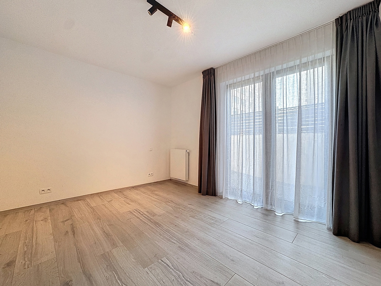 Pièce Lumineuse avec Baie Vitrée – You Real Estate Agency Pièce lumineuse d’appartement avec parquet, grande baie vitrée et rideaux.