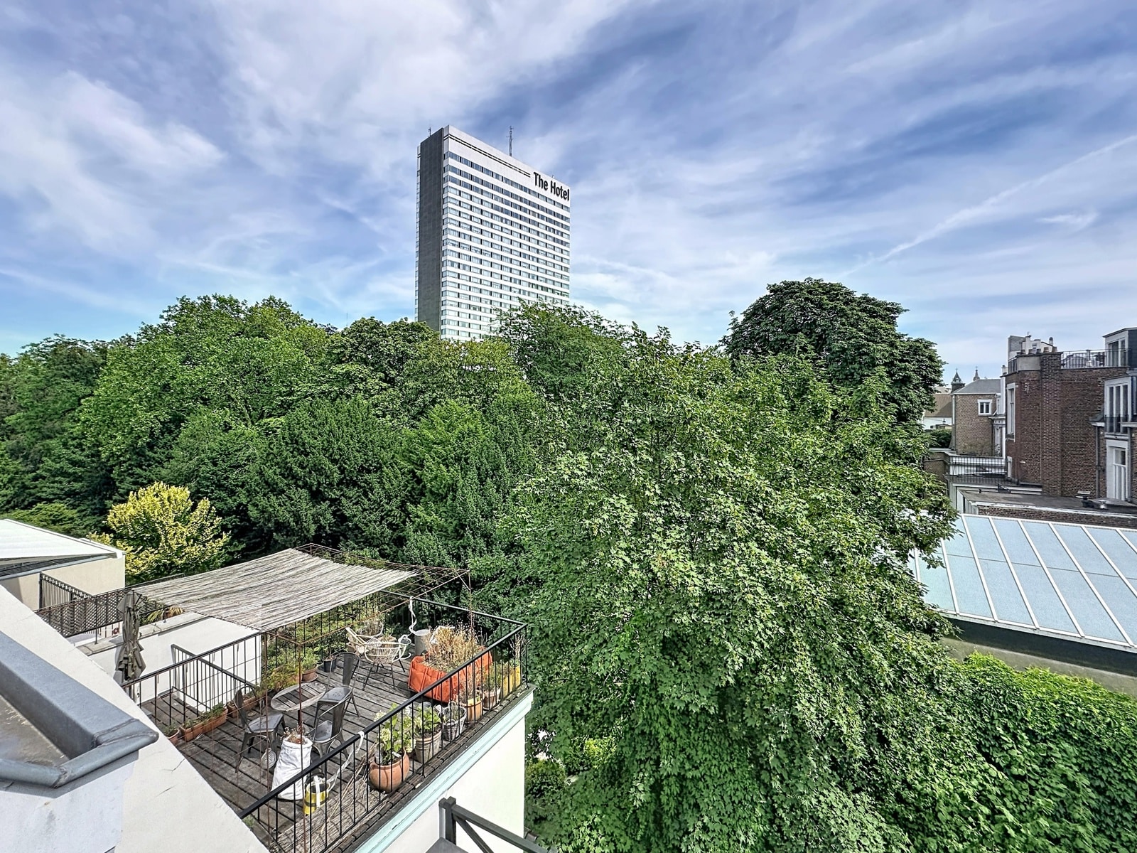 Terrasse Urbaine Avec Vue Arborée – You Real Estate Agency Terrasse d’appartement avec vue urbaine, verdure et tour hôtelière en arrière-plan.