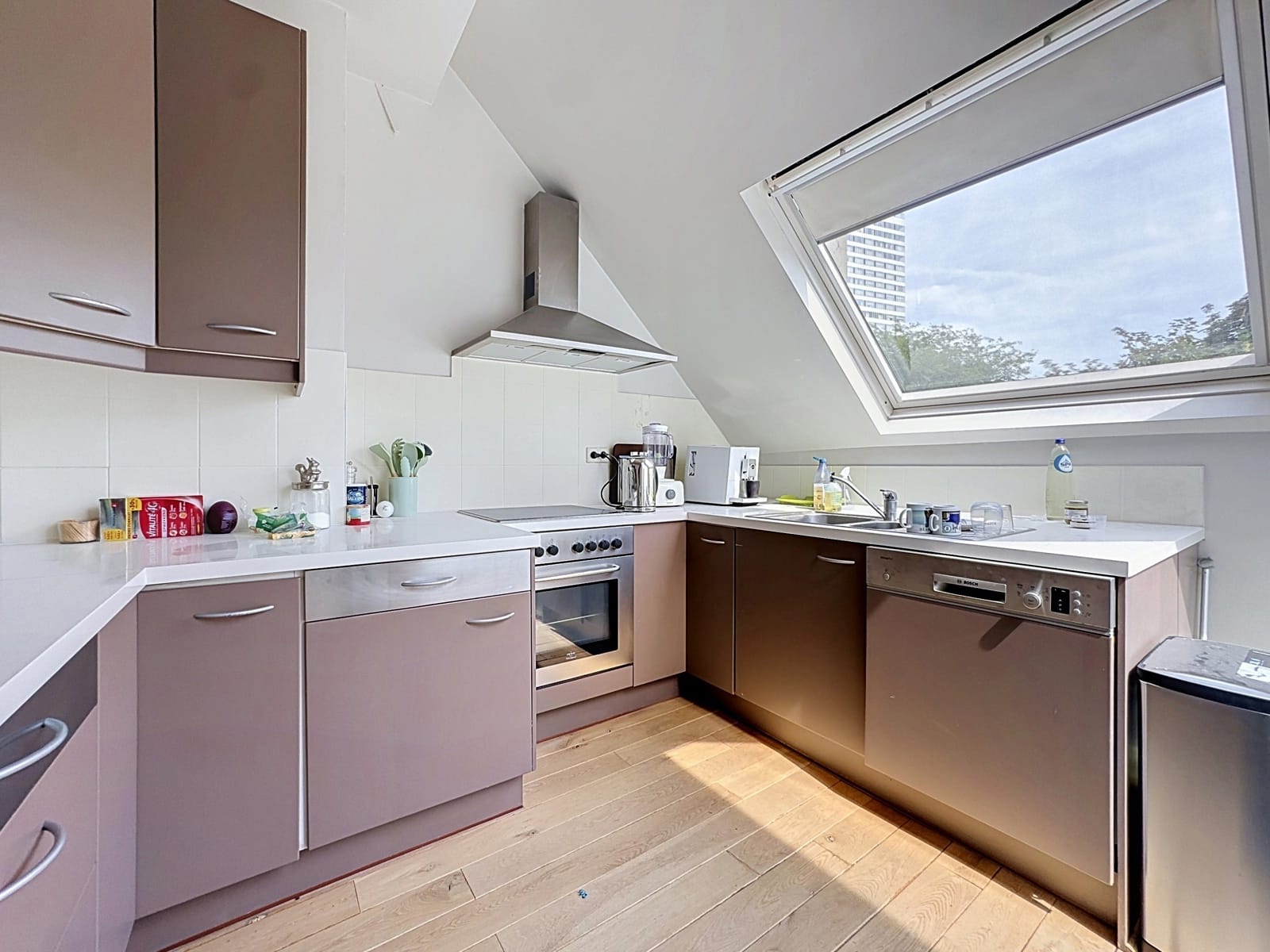 whise photo 6 Modern L-shaped kitchen under a slanted ceiling with a large skylight, brown cabinets, and stainless-steel appliances including oven and dishwasher.