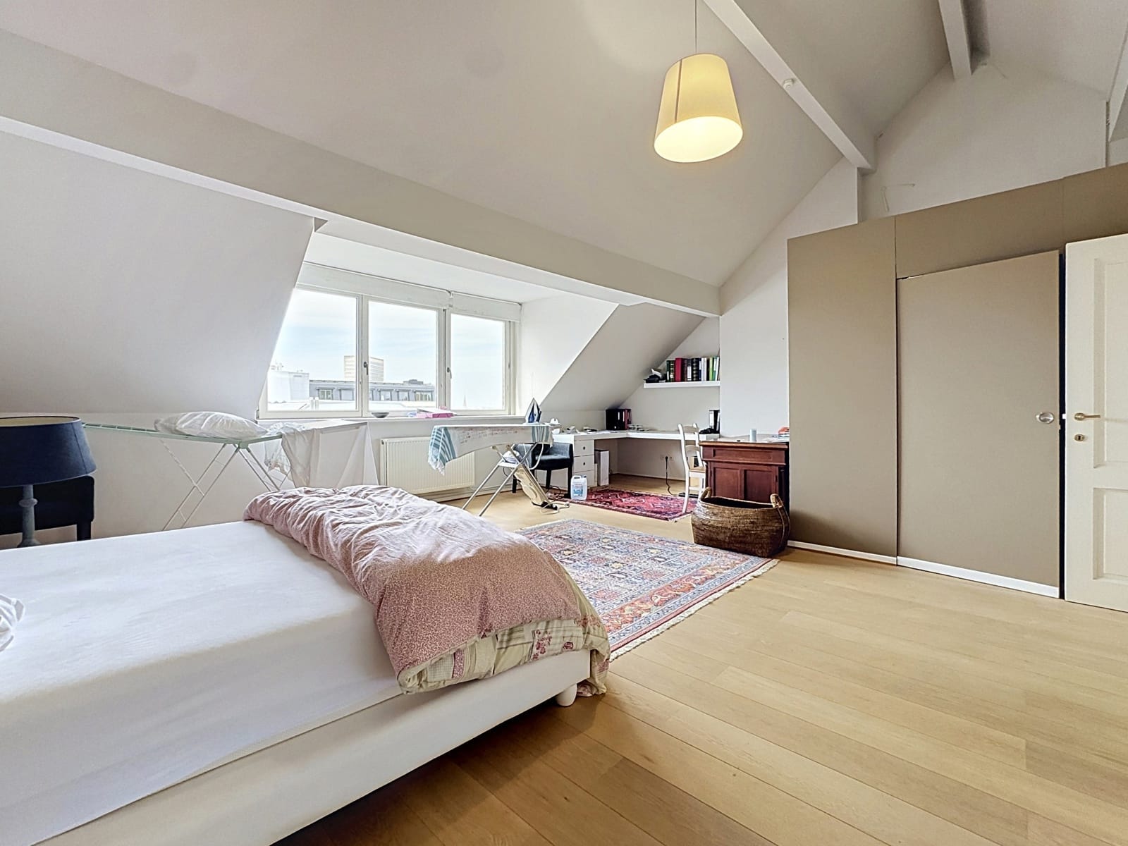 whise photo 7 Bright attic bedroom with a slanted ceiling, large window, and a bed in the foreground; a desk, shelves, and wardrobe along the wall.