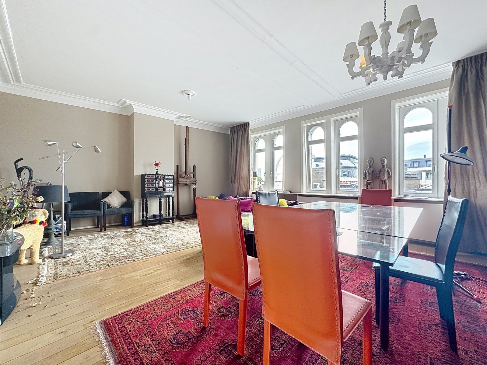 Salon et Salle à Manger Lumineux – You Real Estate Agency Bright living and dining room with a glass dining table and orange chairs on a red Persian rug; large arched-windowed wall and chandelier above.