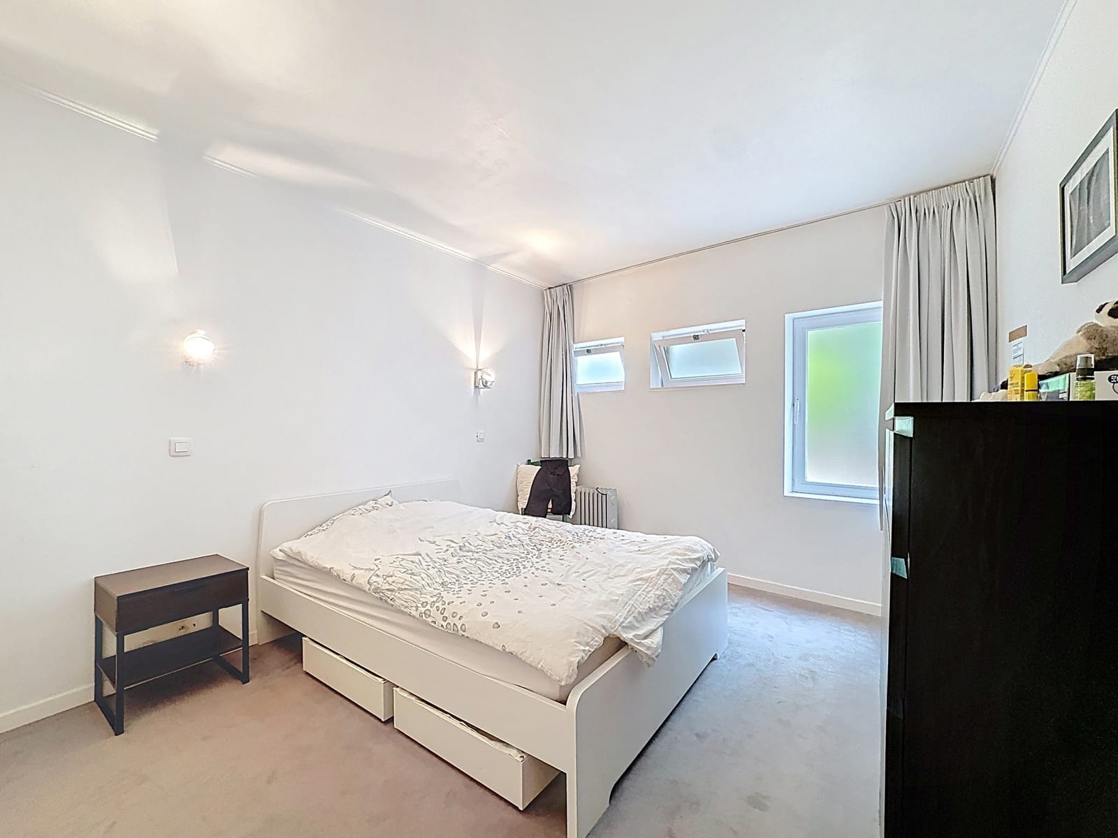 whise photo 7 A bright, minimalist bedroom with a white bed, nightstand, and dresser; soft wall sconces, a beige carpet, and light gray curtains next to small frosted windows.