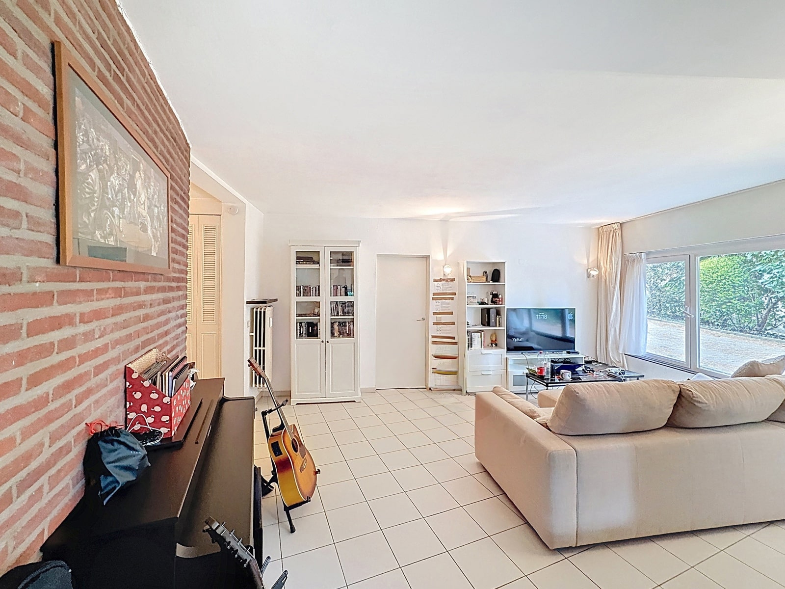 whise photo 3 A bright living room with a brick accent wall, a piano and guitars, white display cabinets, a beige sofa, and a large window that lets in light.