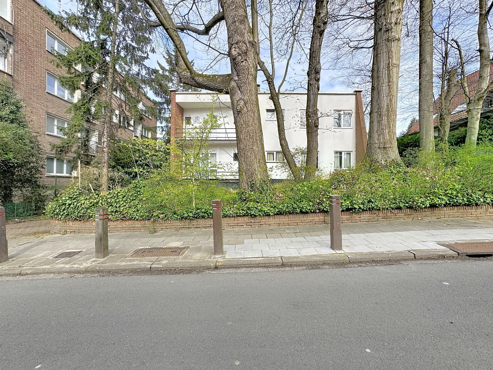whise photo 9 A white apartment building visible behind several tall trees and a dense hedge, along a paved sidewalk with bollards in the foreground.