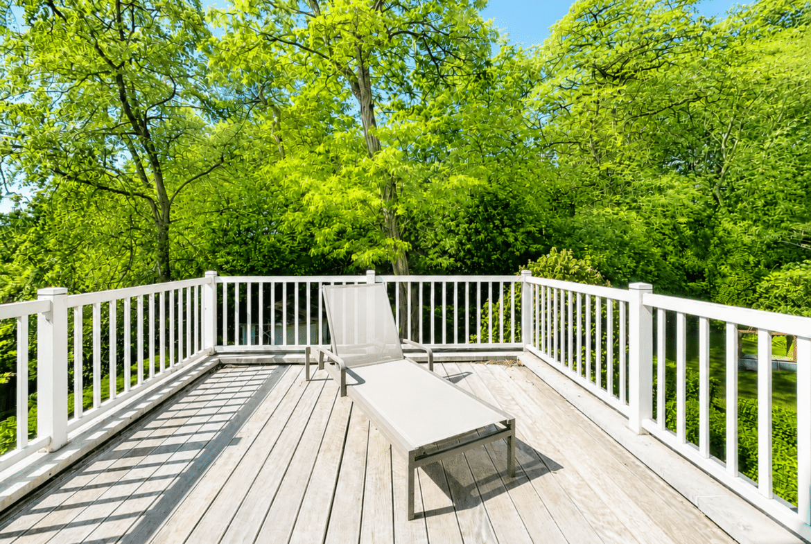 Terrasse en bois ensoleillée avec transat, entourée d’arbres verdoyants.