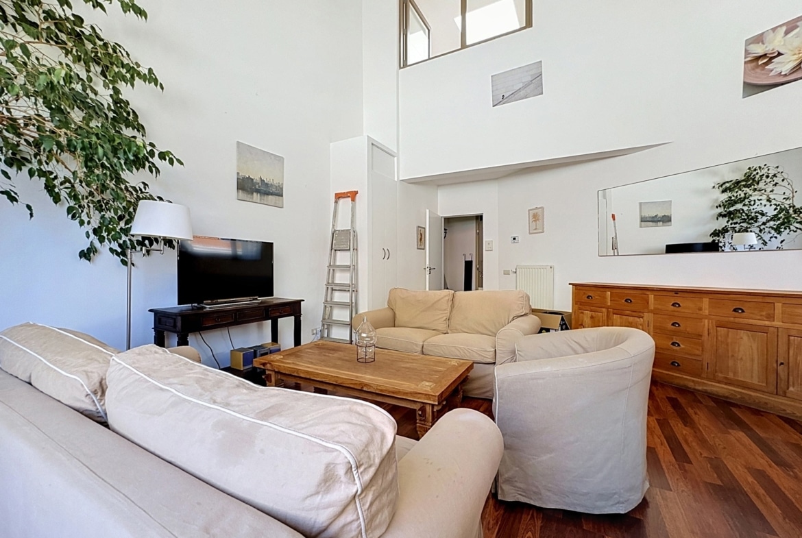Salon lumineux avec hauts plafonds, mobilier beige et parquet en bois.