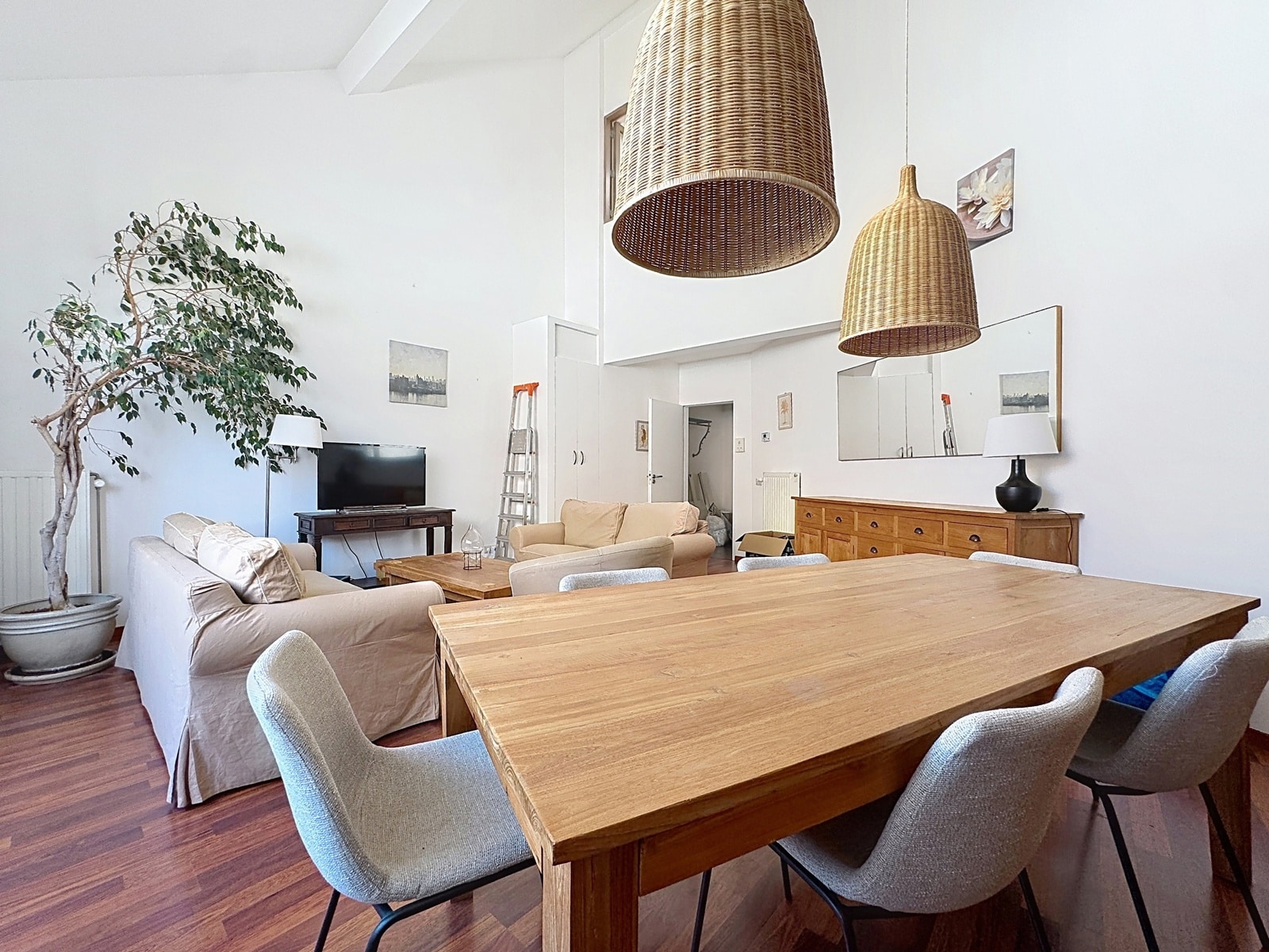 whise photo 5 Open living and dining area with a large wooden table, beige sofas, grey chairs, and two oversized wicker pendant lights overhead.