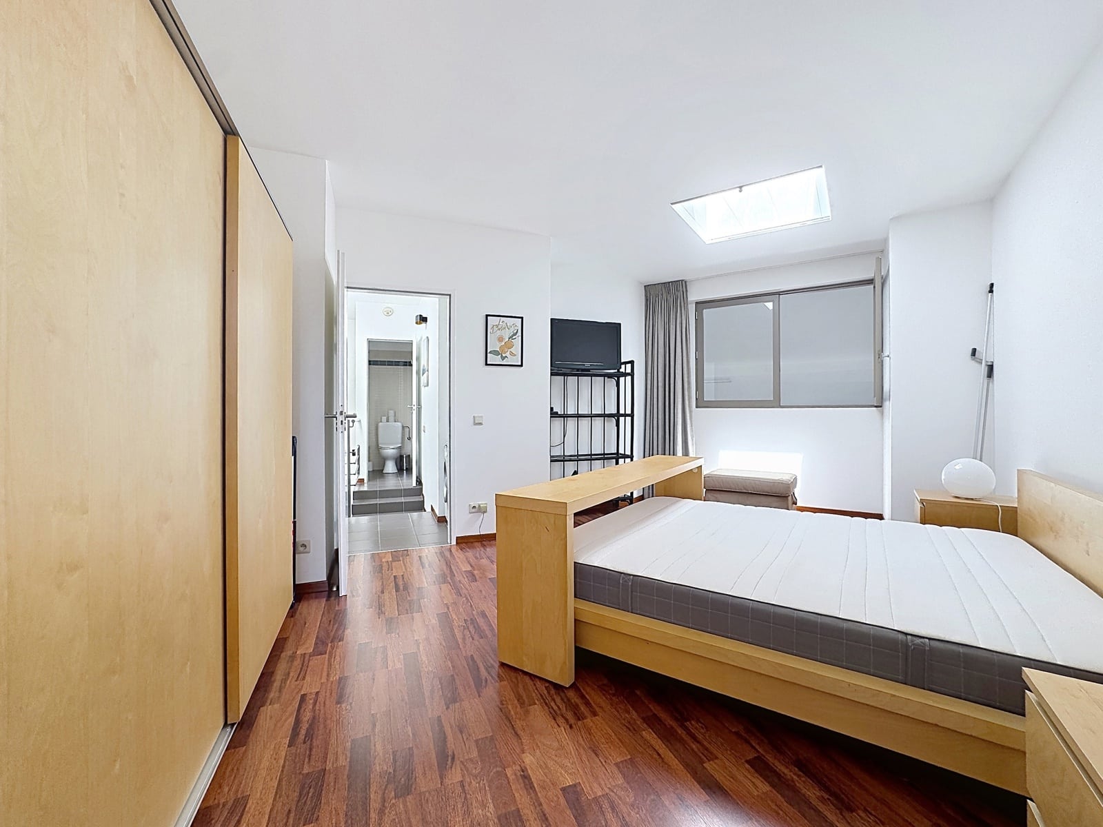 whise photo 11 Bright minimalist bedroom with a wooden bed, large wardrobe, TV on a metal rack, and a doorway to a tiled bathroom visible in the hall area.
