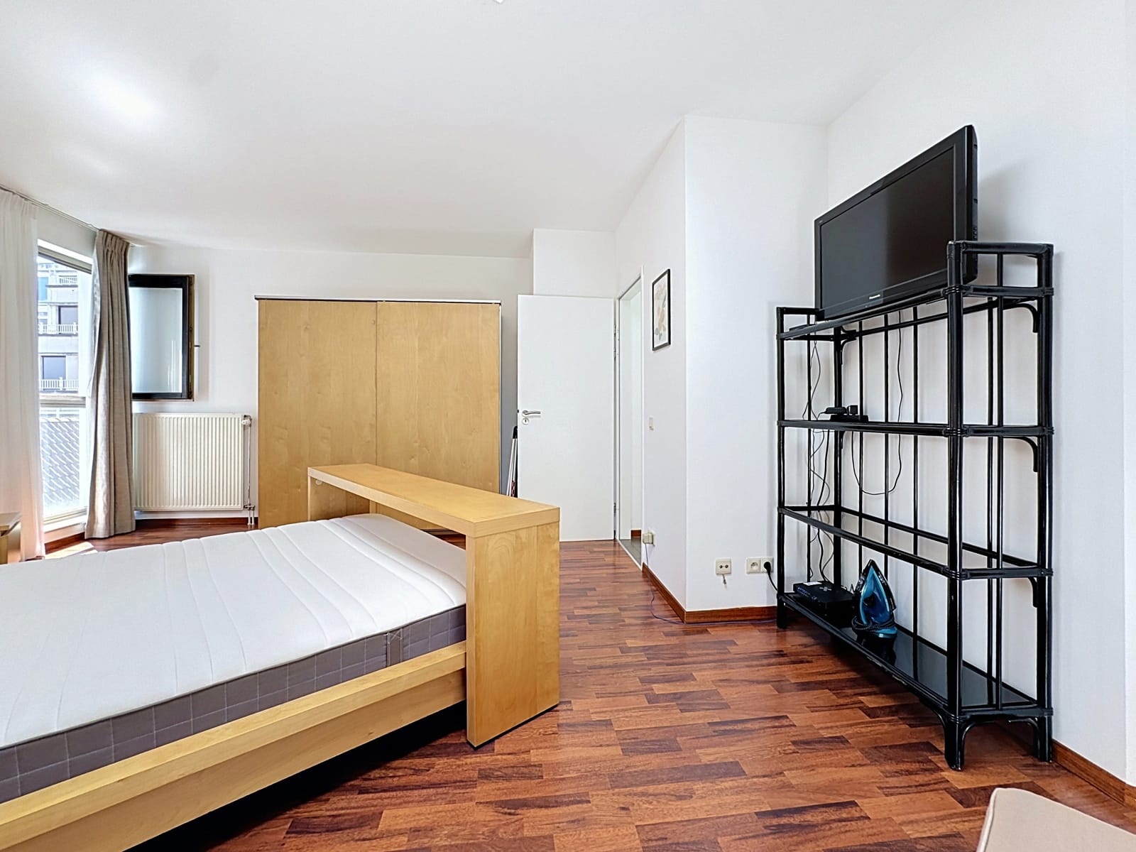 Chambre Lumineuse avec Parquet – You Real Estate Agency Chambre lumineuse d’appartement avec parquet, lit en bois, armoire et étagère TV.