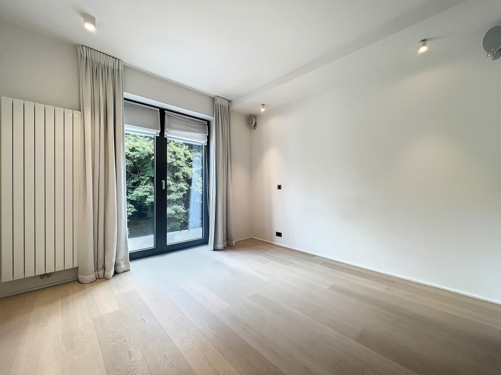 whise photo 8 Empty modern room with light wood flooring, white walls, and a large glass door opening to greenery outside, framed by gray curtains.