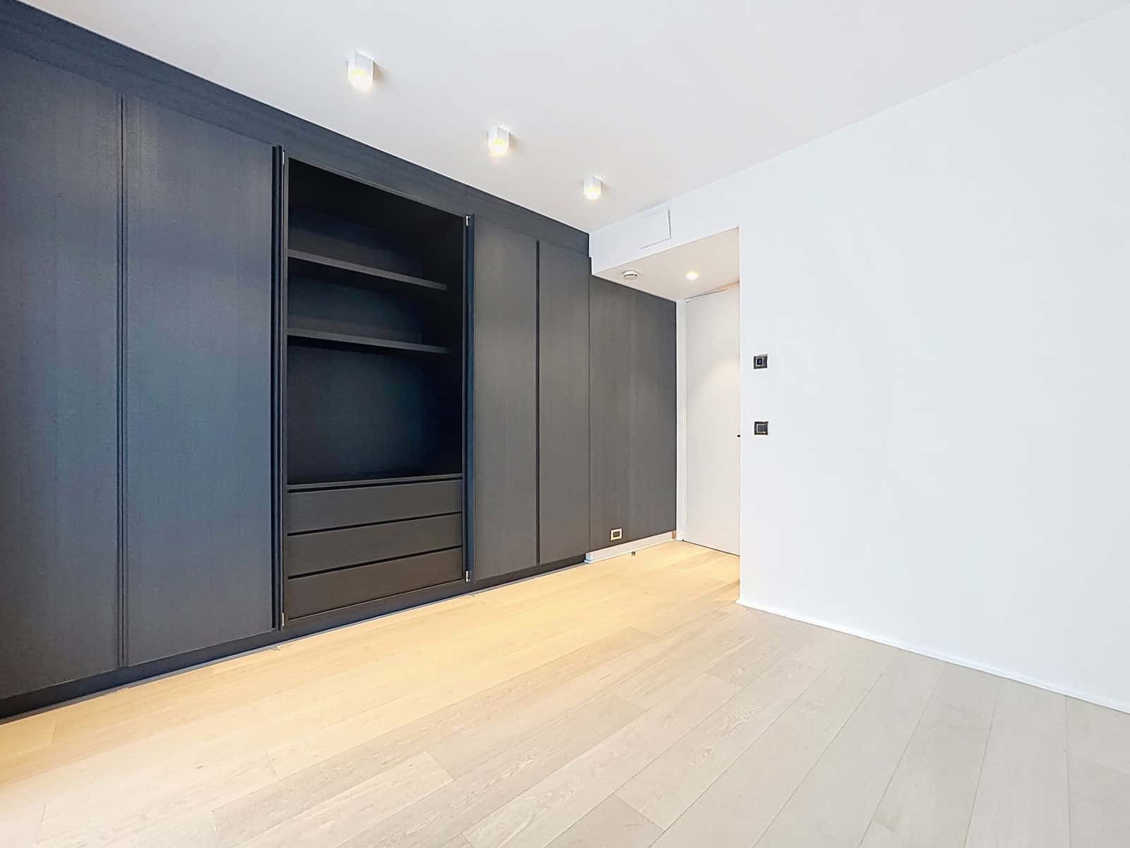 Chambre Moderne avec Dressing Intégré – You Real Estate Agency Modern dark wooden wardrobe with open shelves and drawers against a white wall in a minimal room with light wood flooring.
