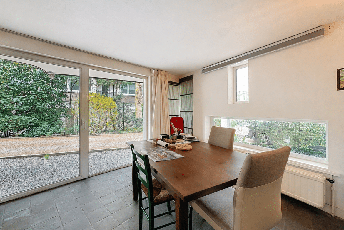 A bright dining room with a wooden table and four chairs, featuring large glass doors that open onto the garden outside.
