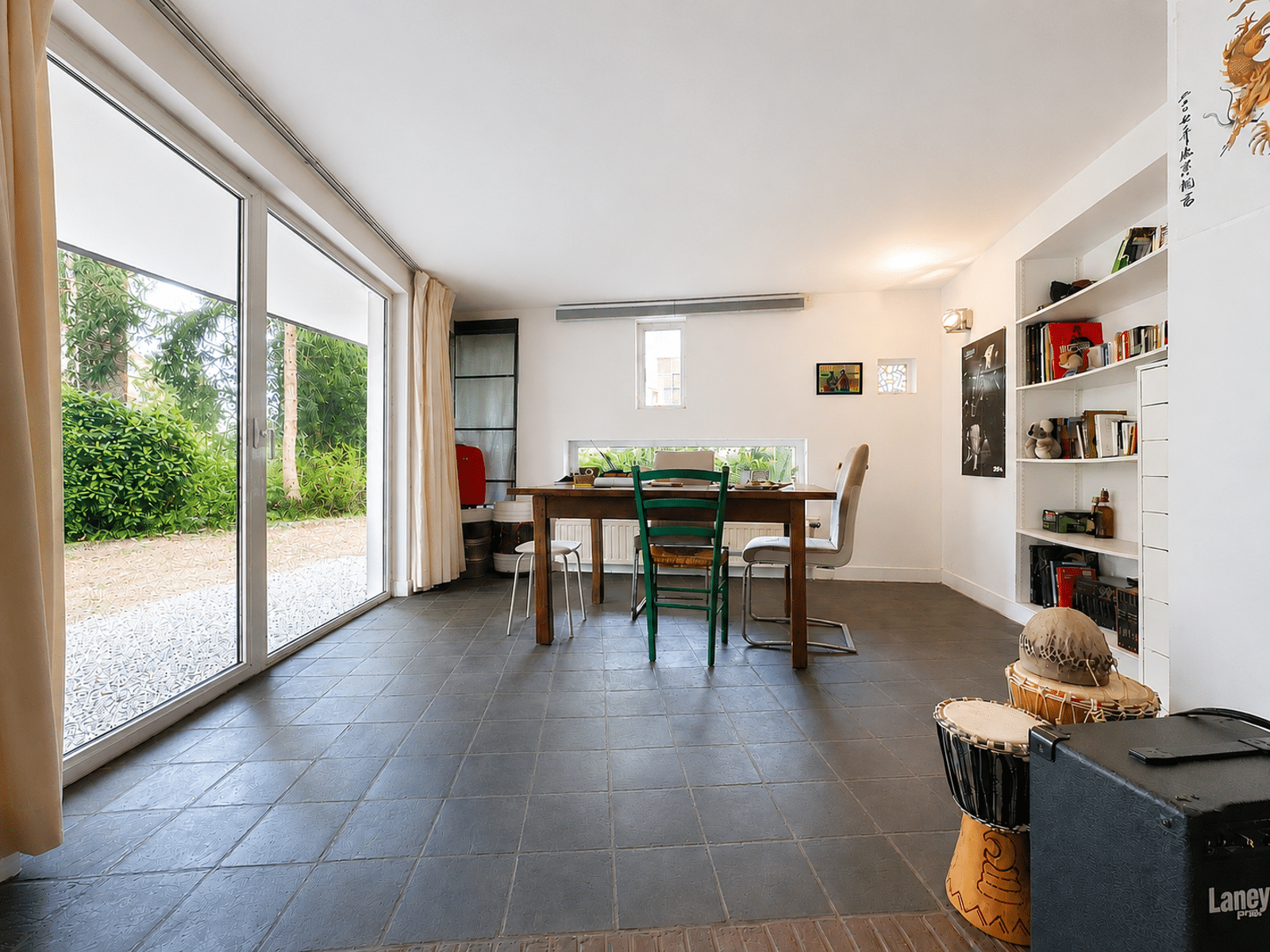 whise photo 2 A bright dining area with a wooden table and a mix of chairs, large sliding glass doors leading to a green garden on the left, white walls, and shelves filled with books on the right, plus a drum and a speaker in the foreground.