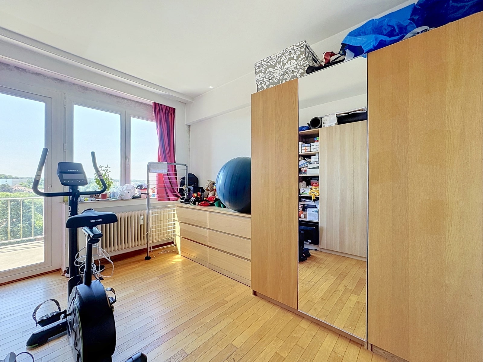 whise photo 8 Home workout area with a stationary bike by a large window, red curtains, and wooden storage units. A blue exercise ball sits on a dresser with clutter nearby.