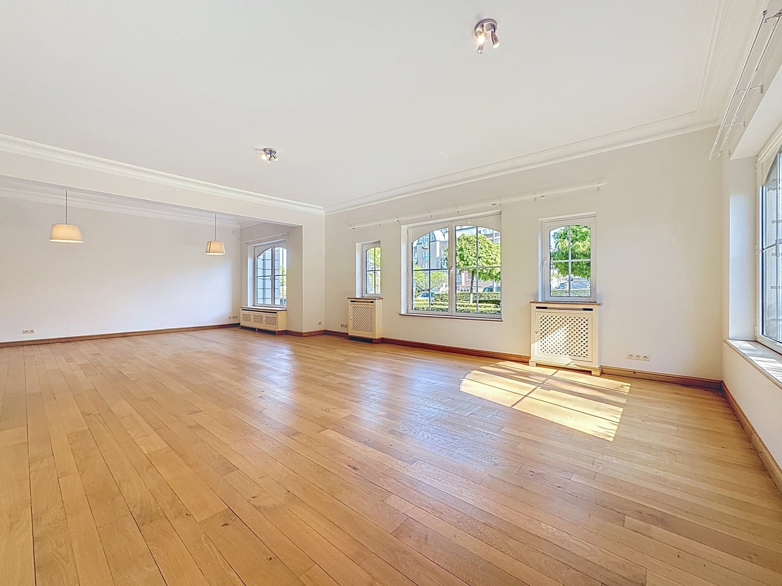 Grand Salon Lumineux avec Parquet – You Real Estate Agency Bright, empty living room with light wood flooring, white walls, tall windows, and radiators beneath the windows, bathed in natural daylight.