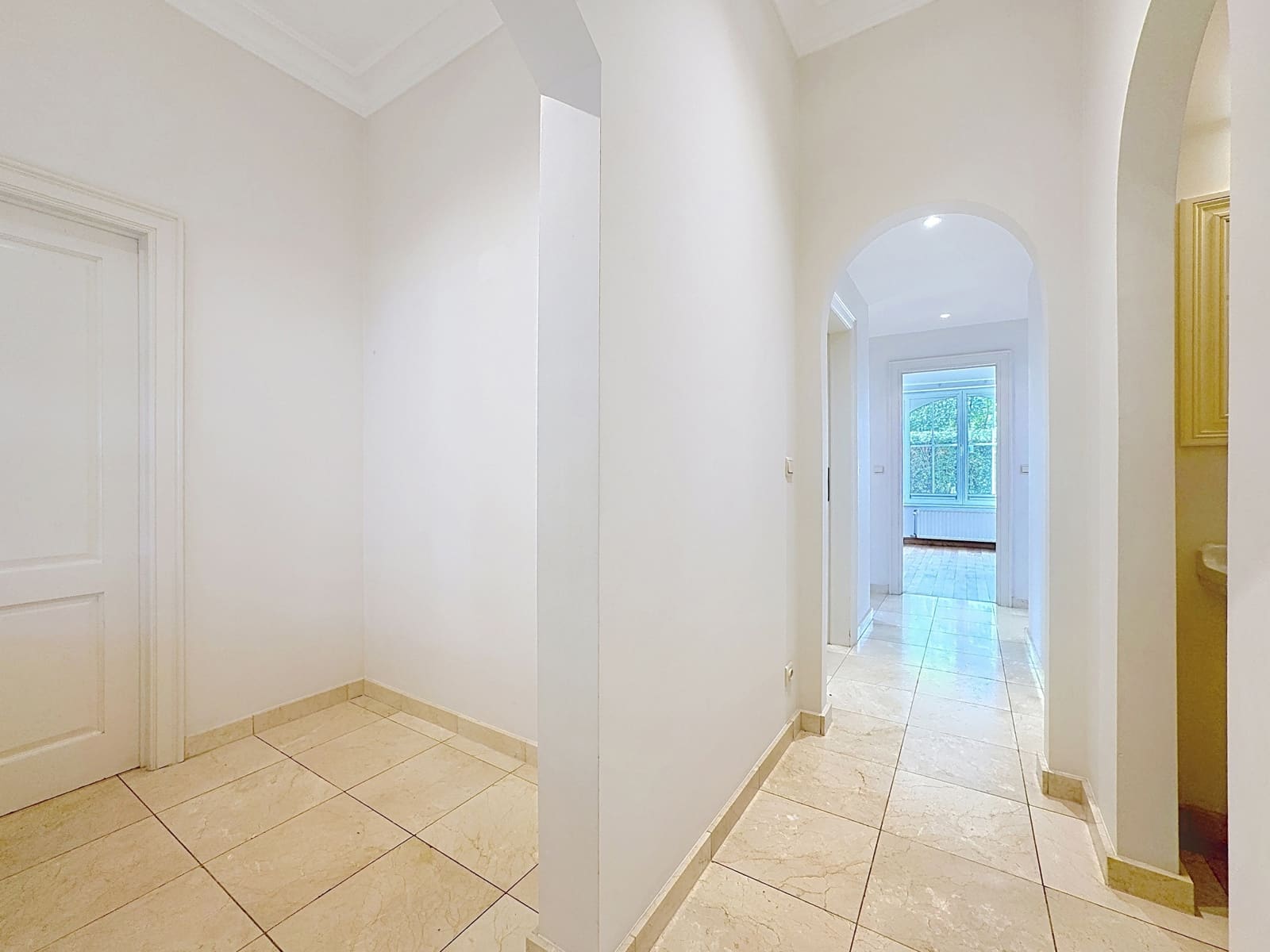 whise photo 8 Bright white hallway with tiled beige floor and a curved arch doorway opening into a sunlit room with a large window at the end.