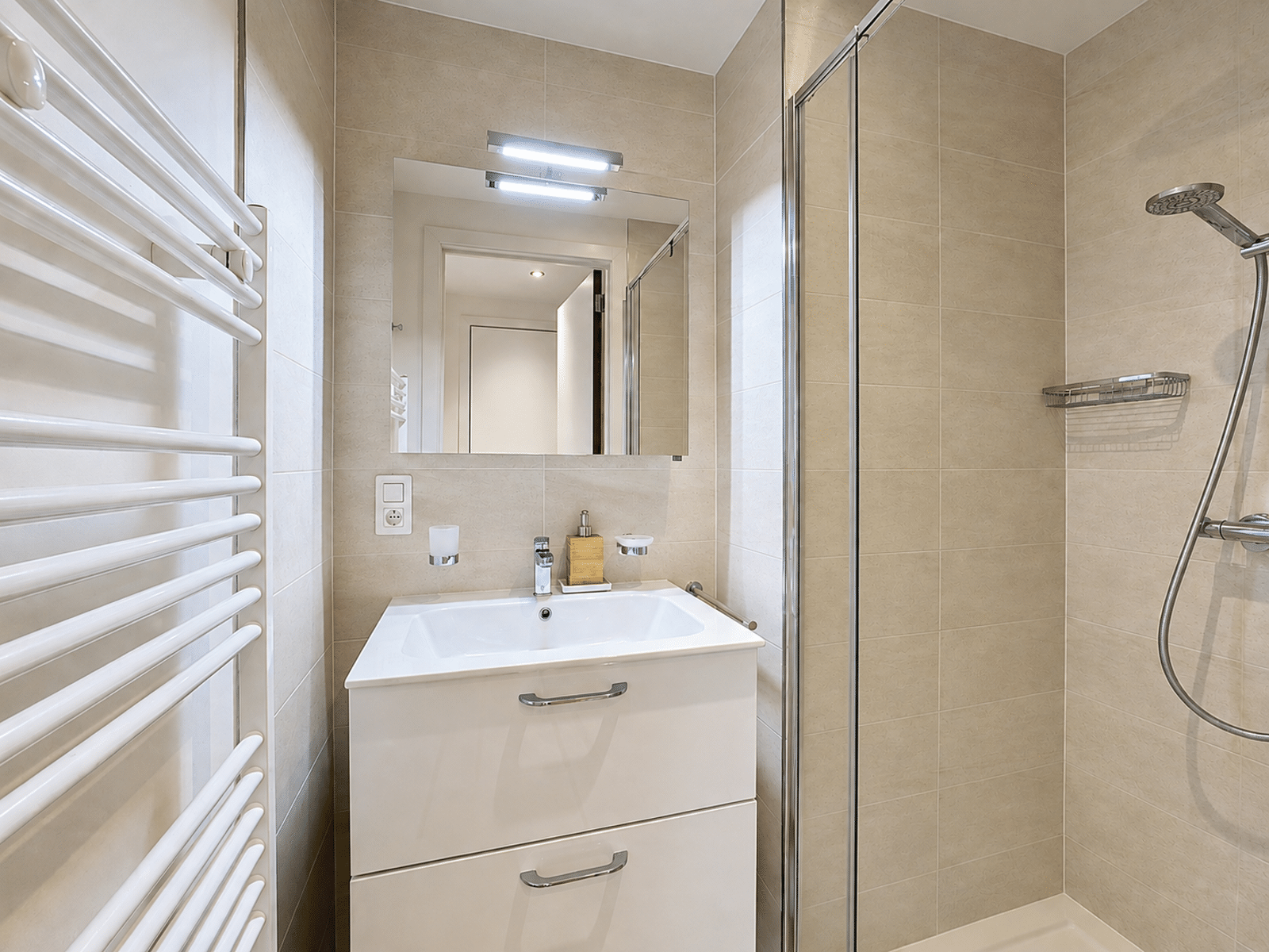 Salle de Bain Moderne avec Douche Vitrée – You Real Estate Agency Modern beige-tiled bathroom with a white vanity, mirror, and glass shower enclosure.