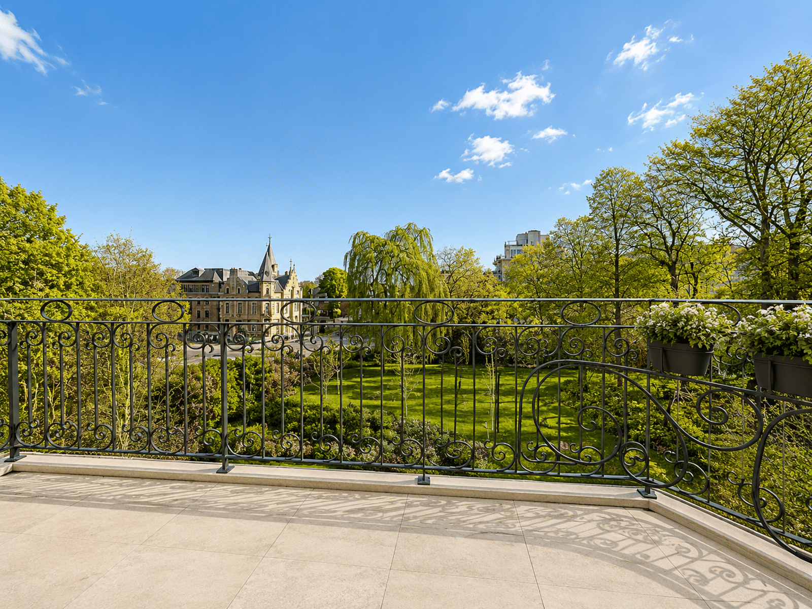 whise photo 15 A view from a balcony overlooking a green lawn and trees, with historic buildings in the distance under a blue sky, featuring an ornamental black railing in the foreground and flower pots on the railing.