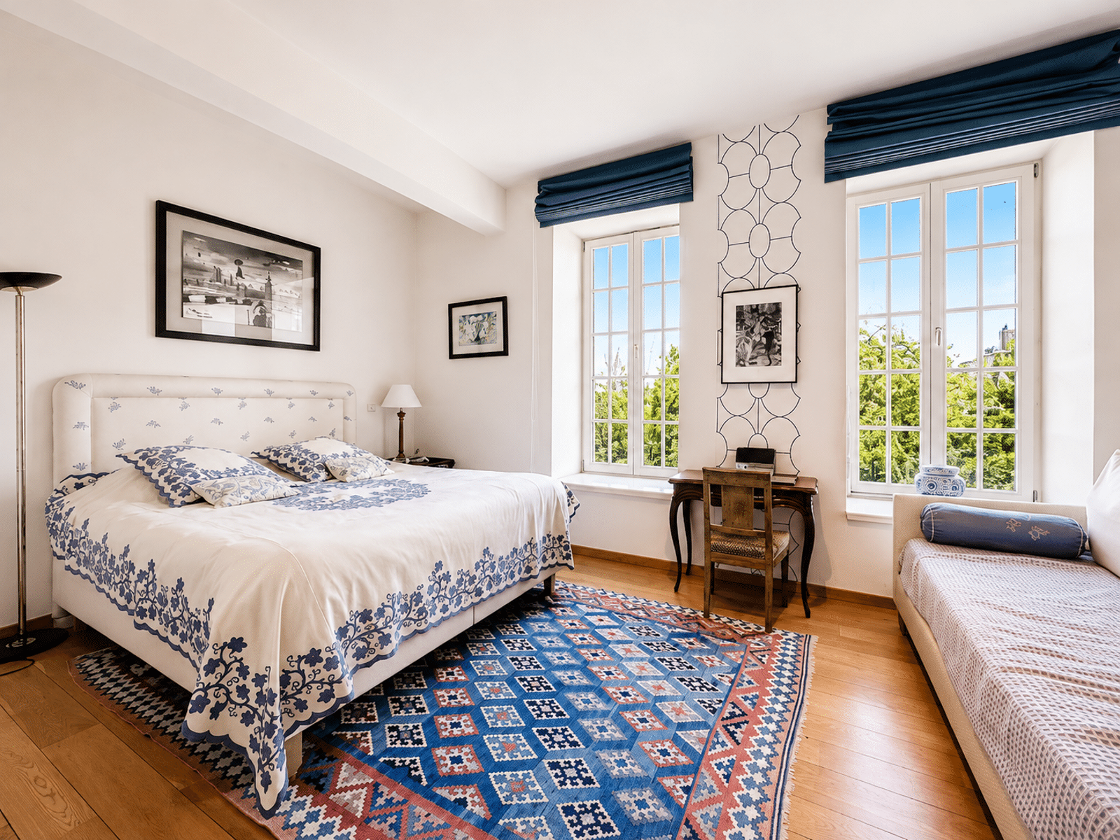 Elegant and Bright Room – You Real Estate Agency An elegant apartment bedroom with a double bed, a desk, hardwood floors, and bright windows.
