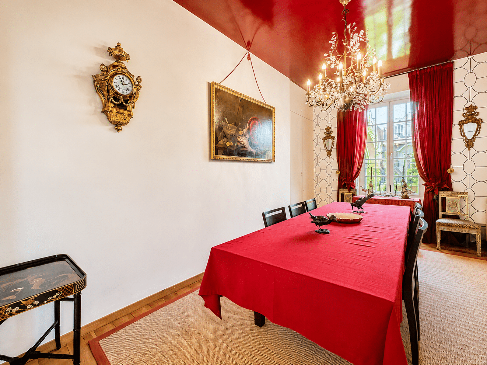 Elegant Dining Room with a Chandelier – You Real Estate Agency An elegant dining room with a red table, a chandelier, and a large window in an apartment.