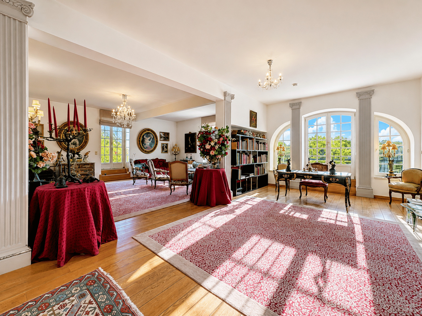 Bright Living Room – You Real Estate Agency A bright living room in a mansion, featuring columns, hardwood floors, chandeliers, and large windows.