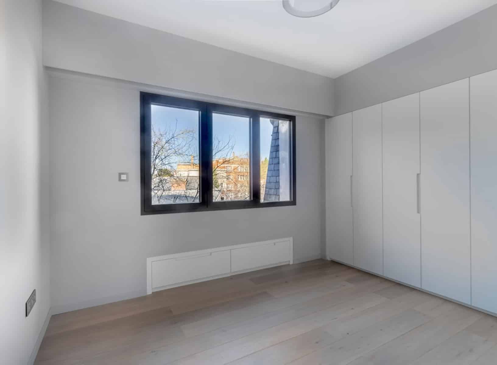whise photo 19 Empty modern room with white built-in wardrobes along the right wall, a large dark-framed window, and light wood floors.