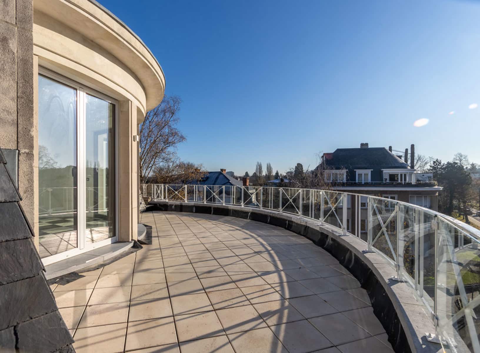 whise photo 5 Curved balcony with glass railing on a sunny building, overlooking rooftops and leafless trees under a blue sky.
