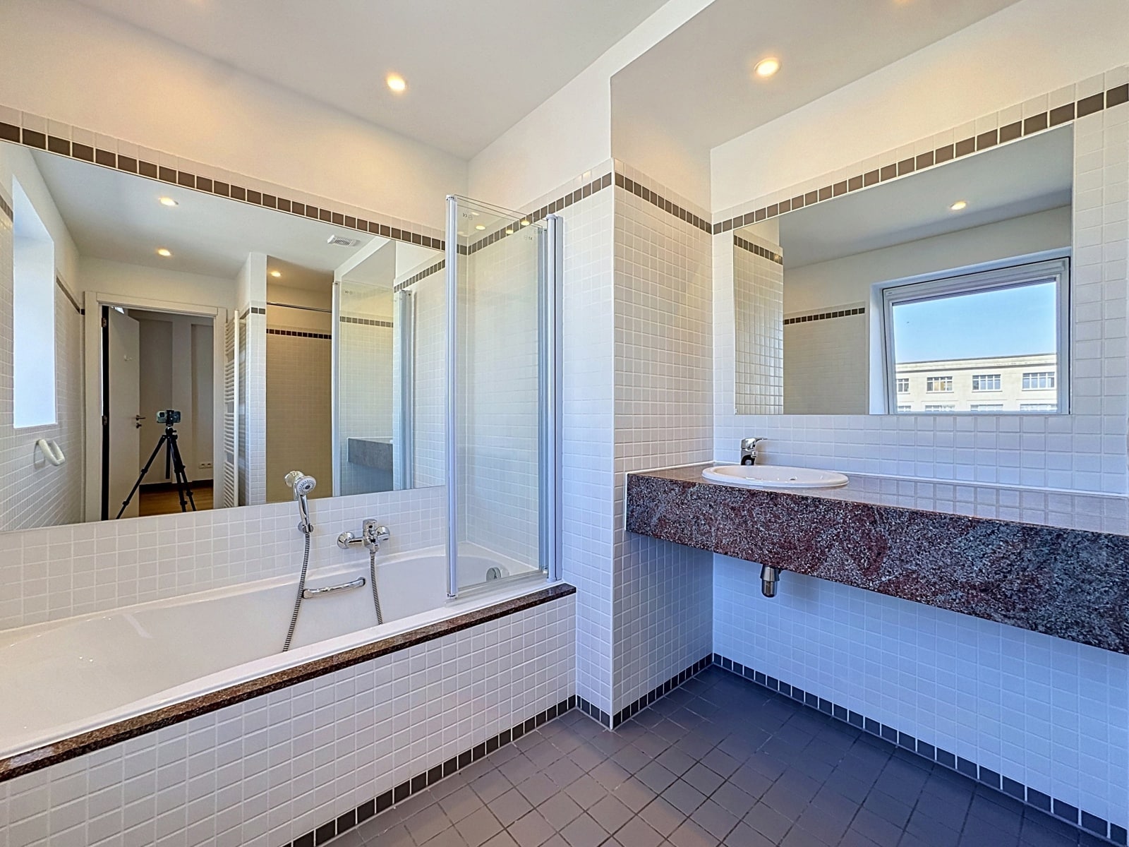 whise photo 7 A bright, modern bathroom with a white-tiled bathtub, a glass shower, a large mirror, and a granite vanity; a camera on a tripod is visible in the doorway.