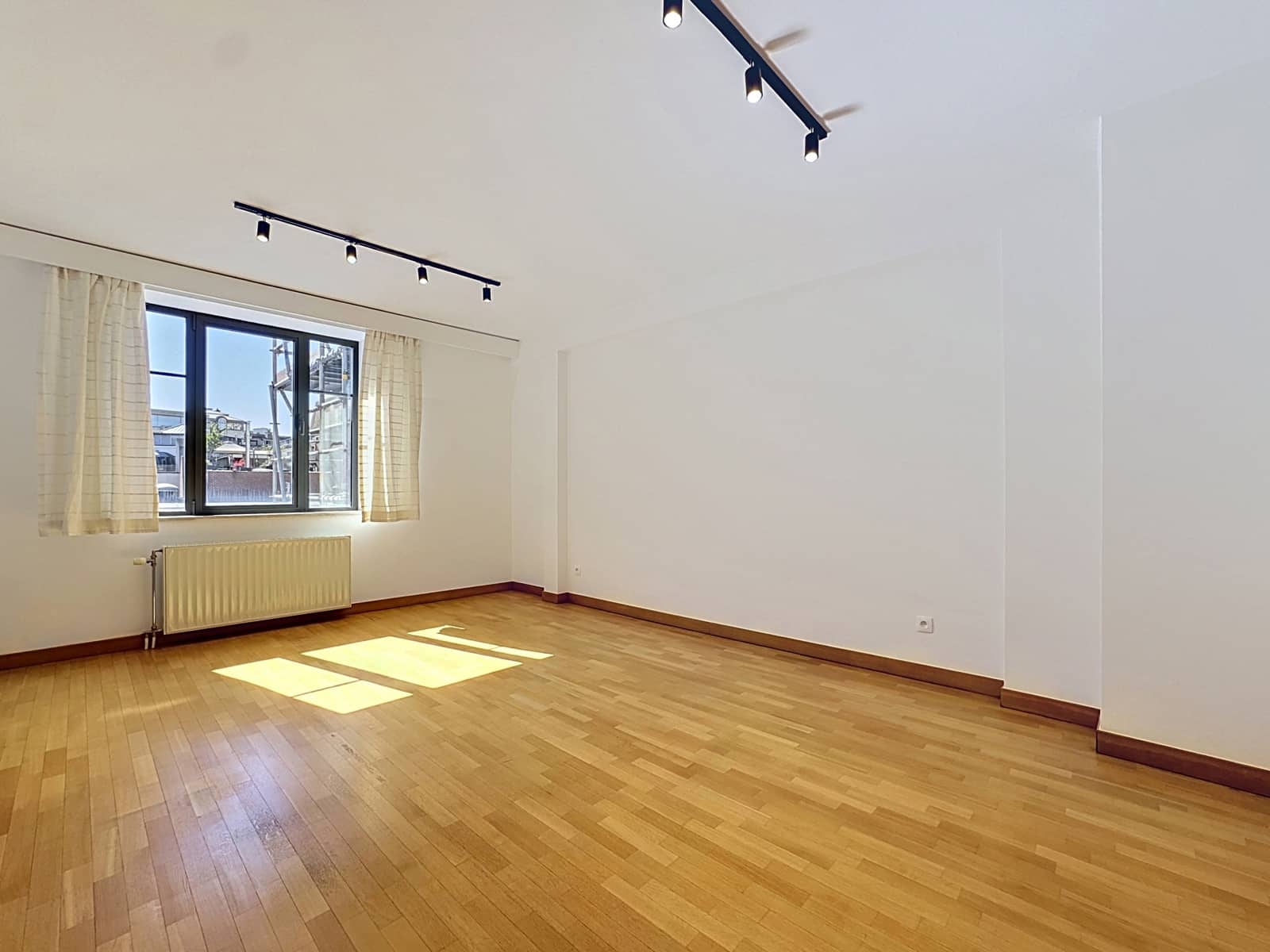 whise photo 2 A sunlit, empty room with light wood floors, a large window with beige curtains, a white radiator beneath the window, and track lighting on the ceiling.