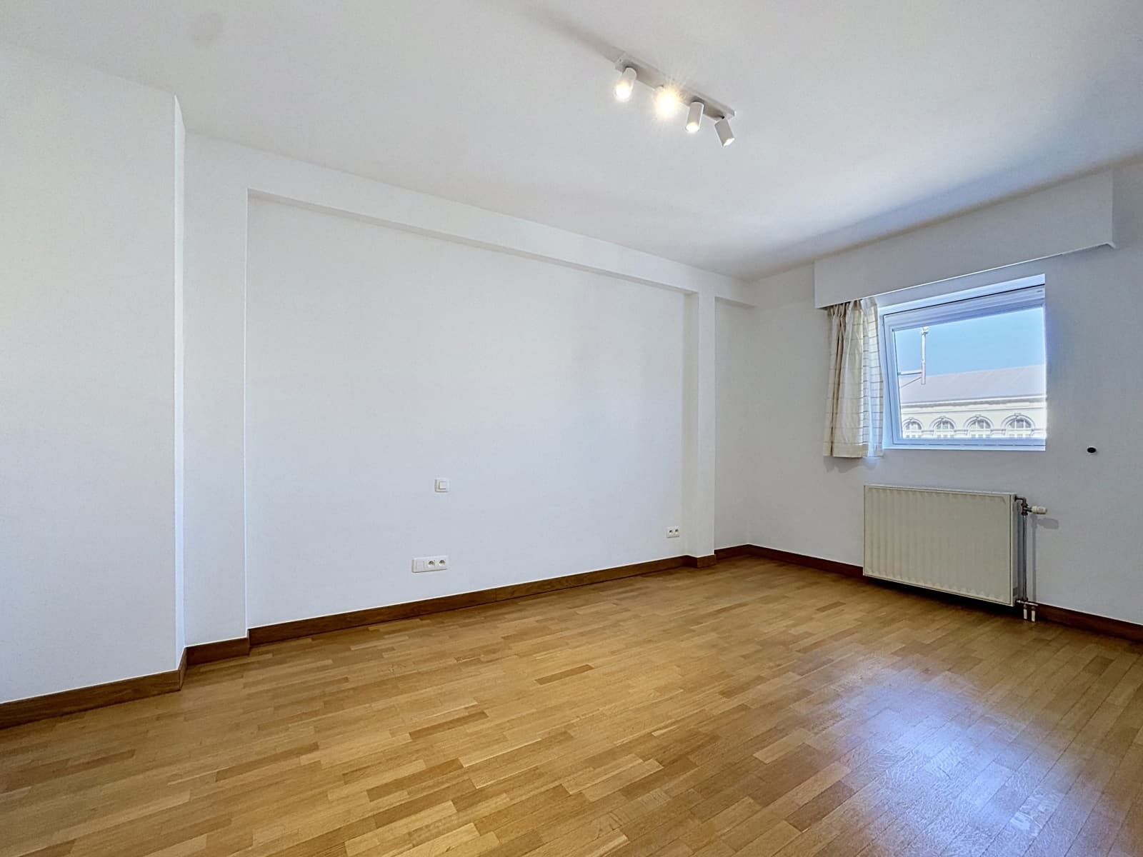 whise photo 5 A bright, empty room with light wood floors, white walls, a window with a beige curtain, and a radiator underneath.