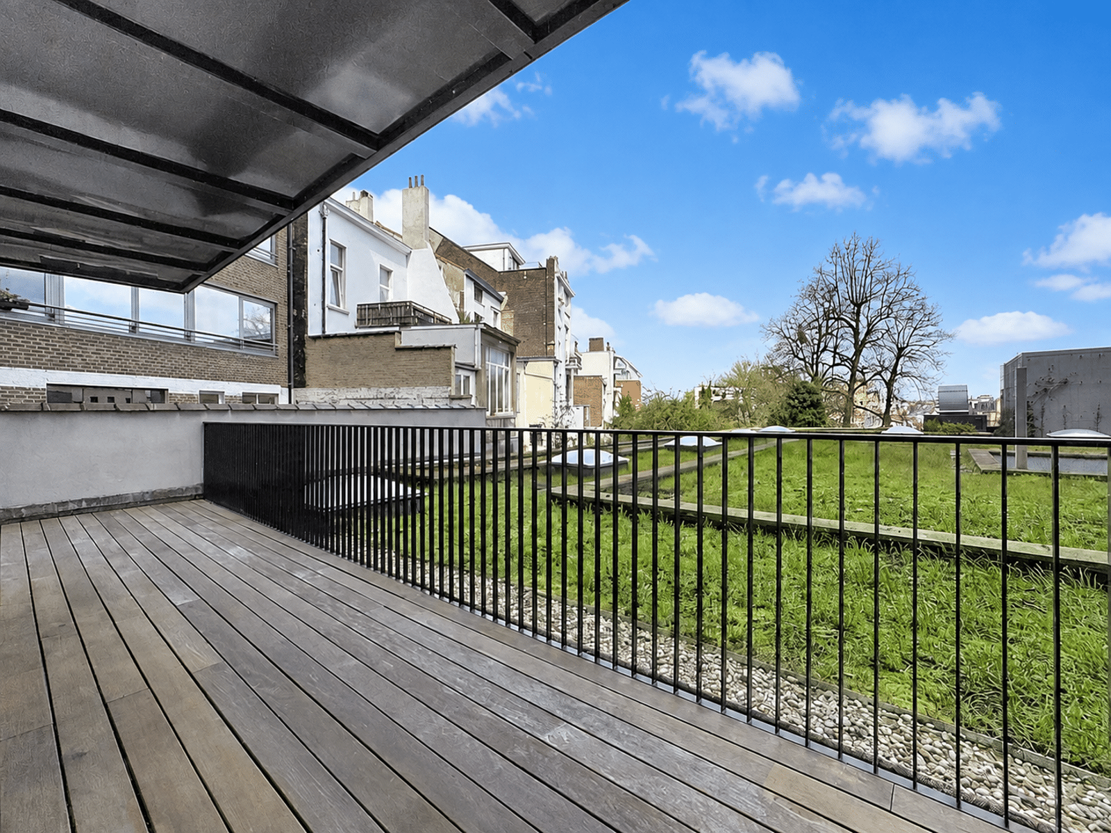 whise photo 4 Wooden balcony with a black railing overlooking a green courtyard and row of townhouses under a bright blue sky.