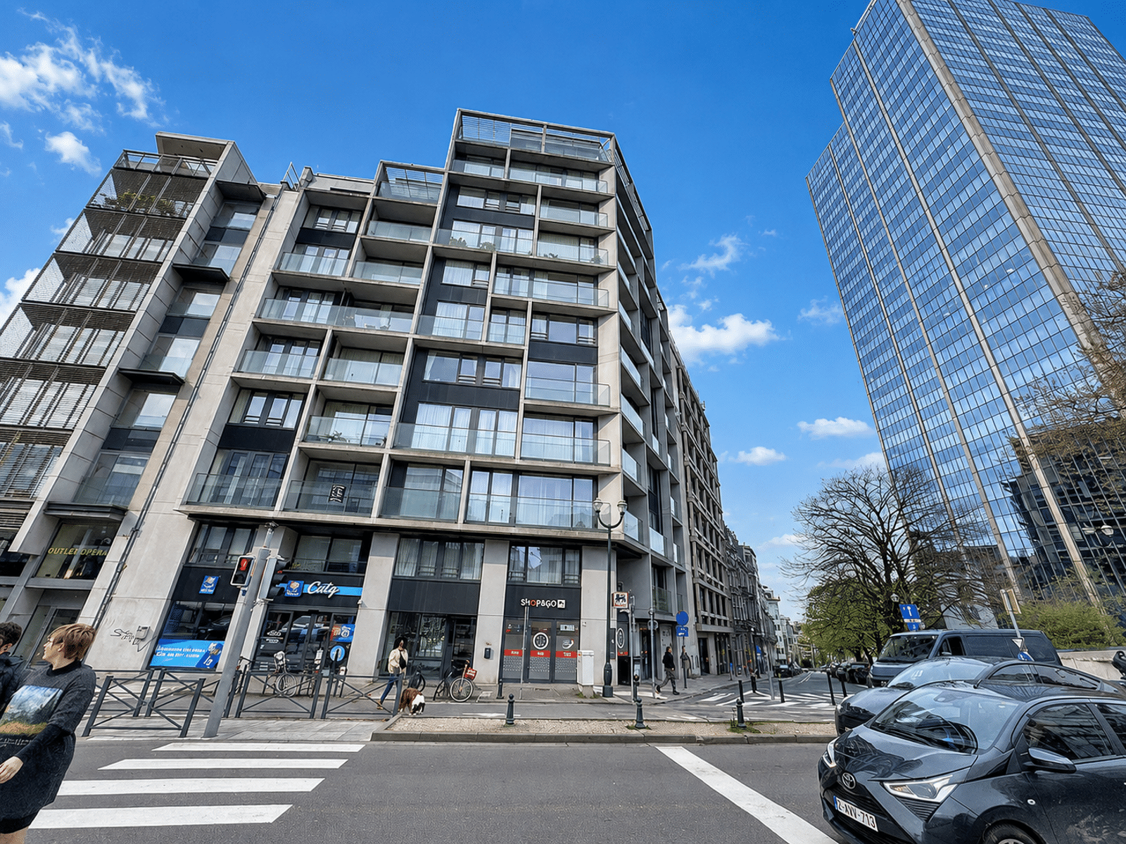 whise photo 11 Urban street with modern multi-story buildings, glass balconies, and ground-floor storefronts under a blue sky.