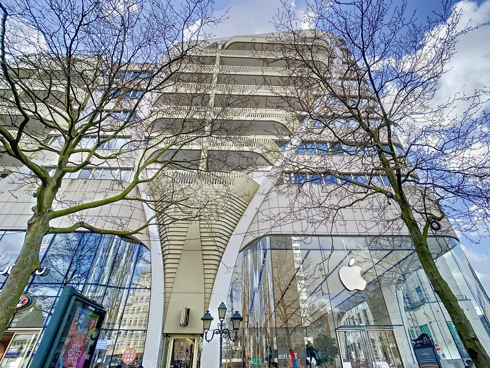 whise photo 16 Low-angle view of a modern building with curved balconies, large glass storefronts, and leafless trees in front.