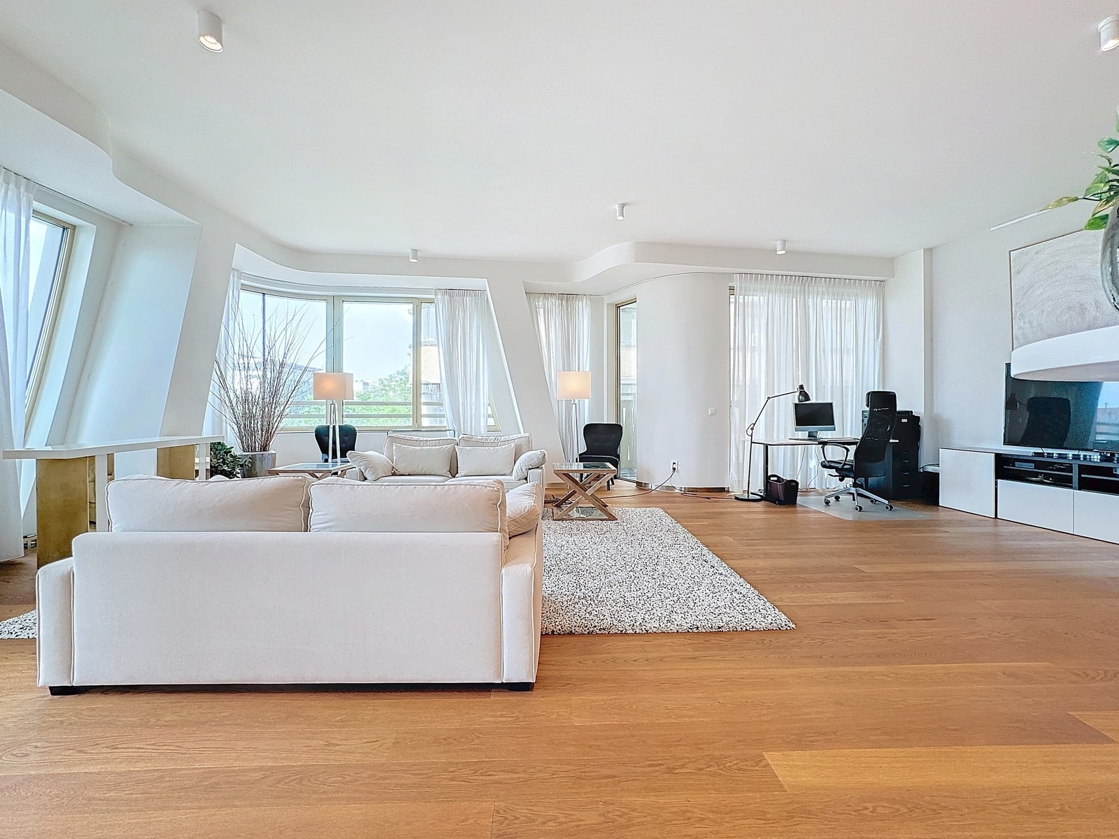 whise photo 6 Bright open-plan living room with white sofas, large windows, and a wooden floor. A cozy seating area faces a rug and coffee table near a desk area to the right.