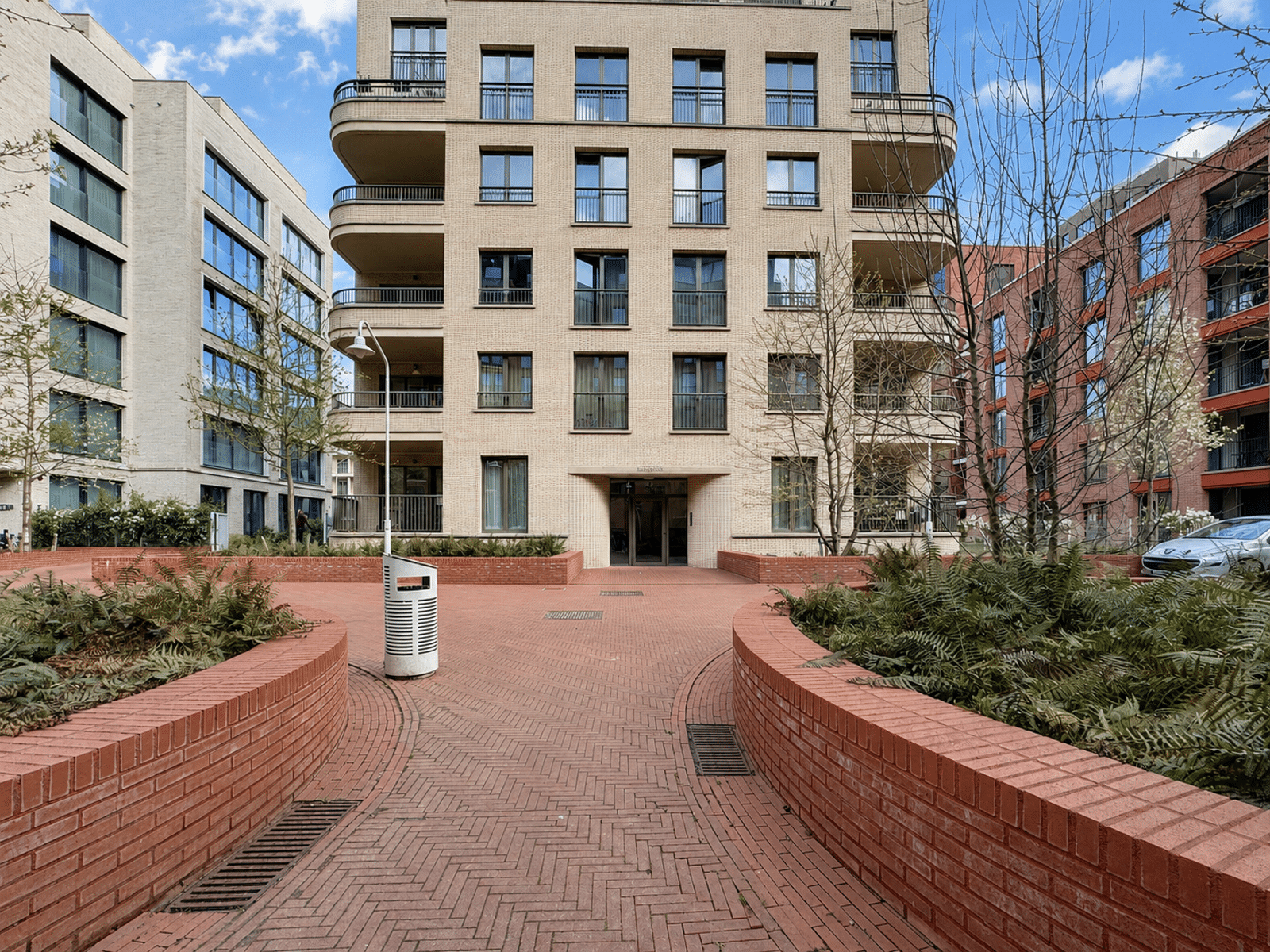 whise photo 12 Brick-paved courtyard with curved brick planters in front of a central beige apartment building with multiple balconies and large windows; blue sky above.