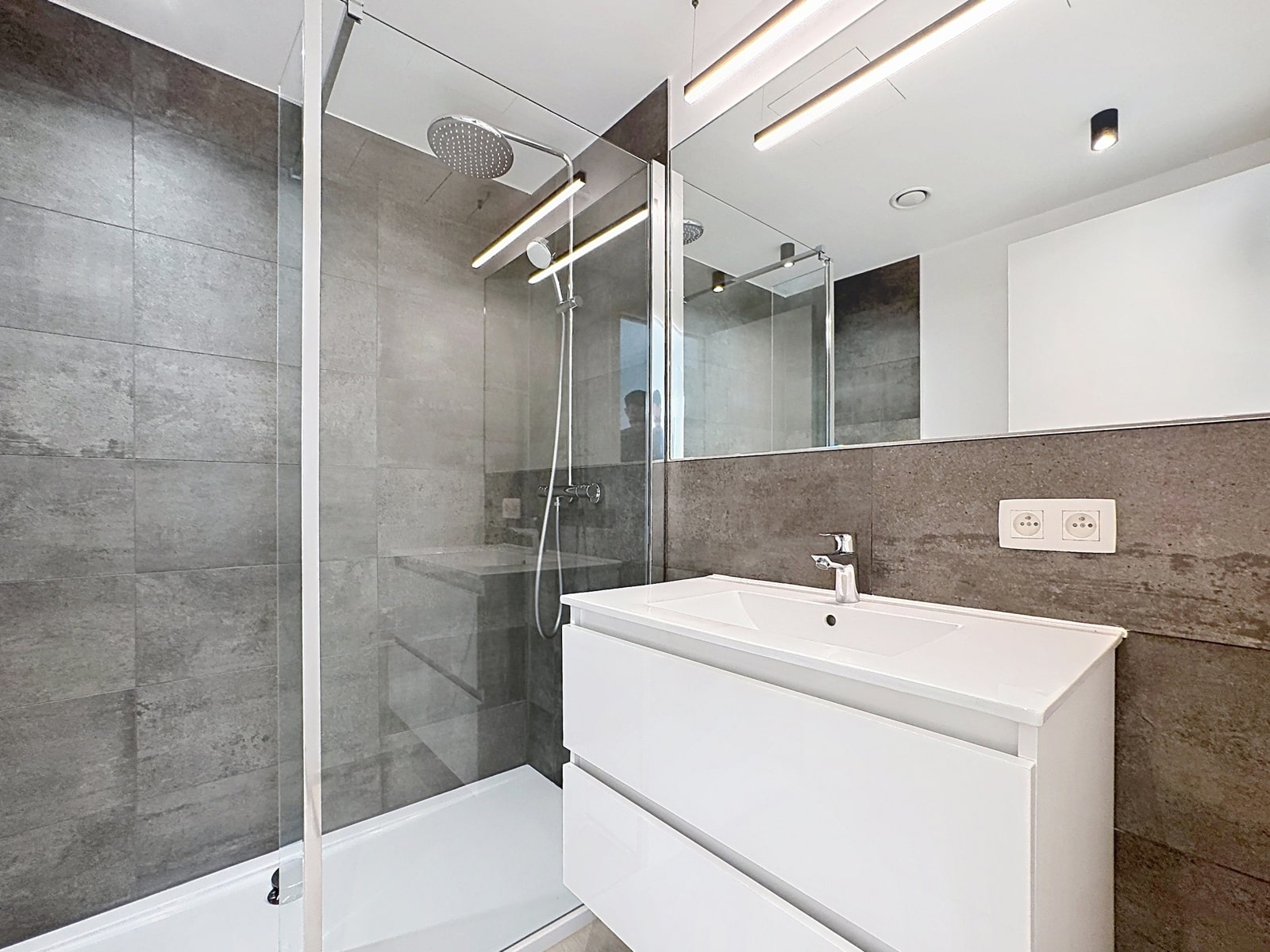whise photo 7 A modern bathroom with a glass shower enclosure, a large rainfall showerhead, and a white vanity set against gray tiled walls.