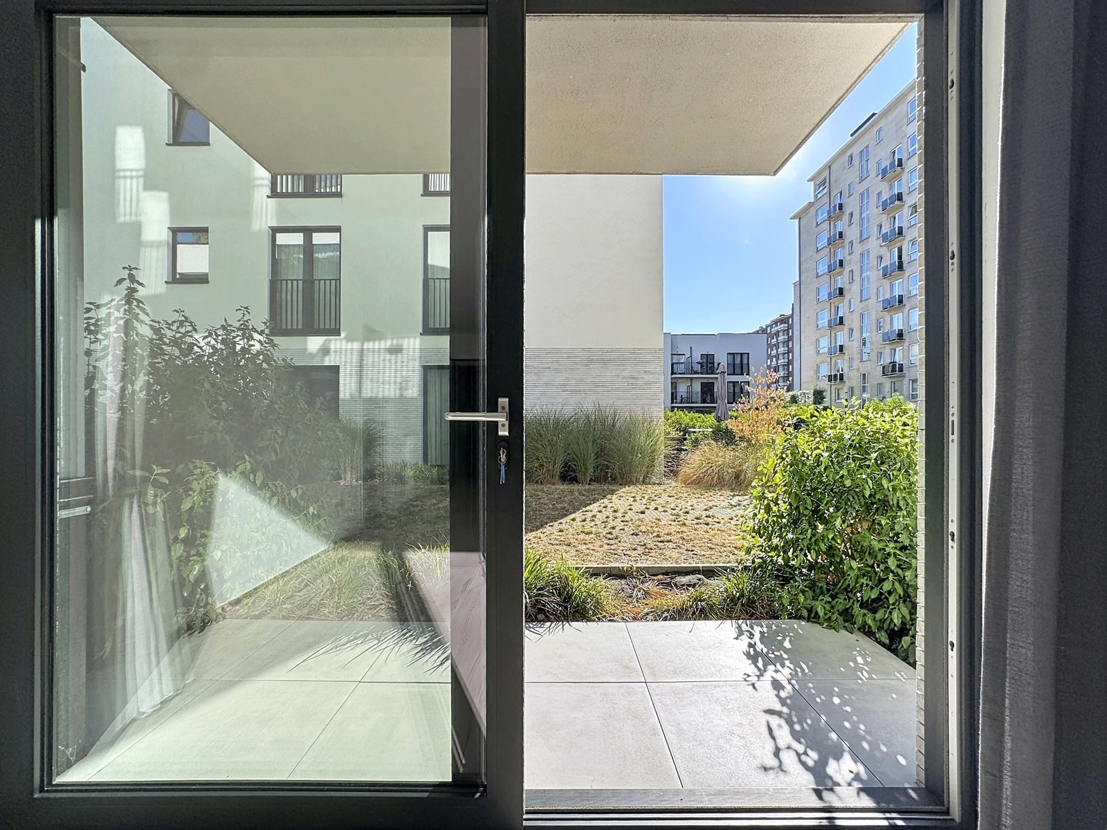 whise photo 8 A view through a glass patio door onto a modern apartment courtyard, with greenery and a blue sky in the distance.