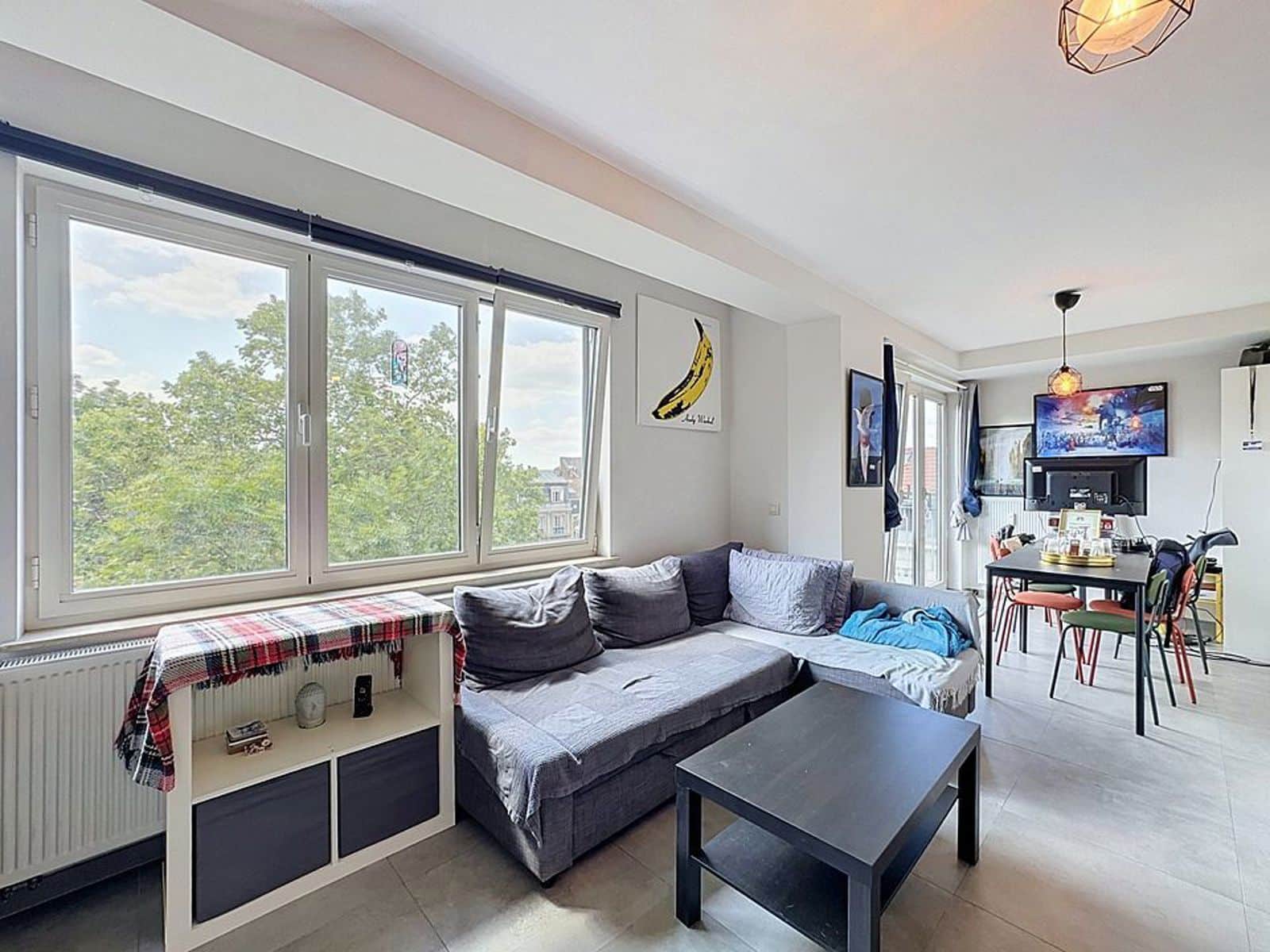 whise photo 2 Bright living room with large windows overlooking trees, a gray sectional sofa, and a black coffee table.