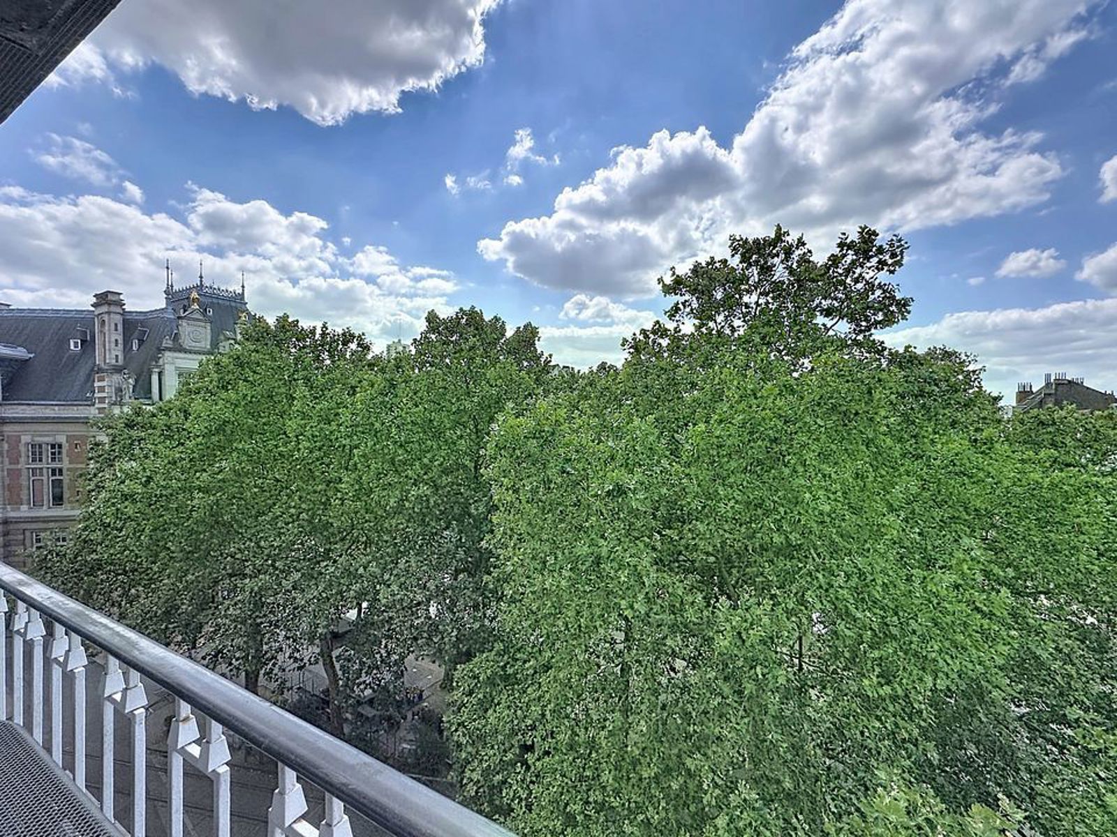 whise photo 8 Balcony view over dense green trees with a historic building on the left and a blue sky with clouds.|Conveys view from a balcony with trees, a historic building to the left, and a sunny sky.
