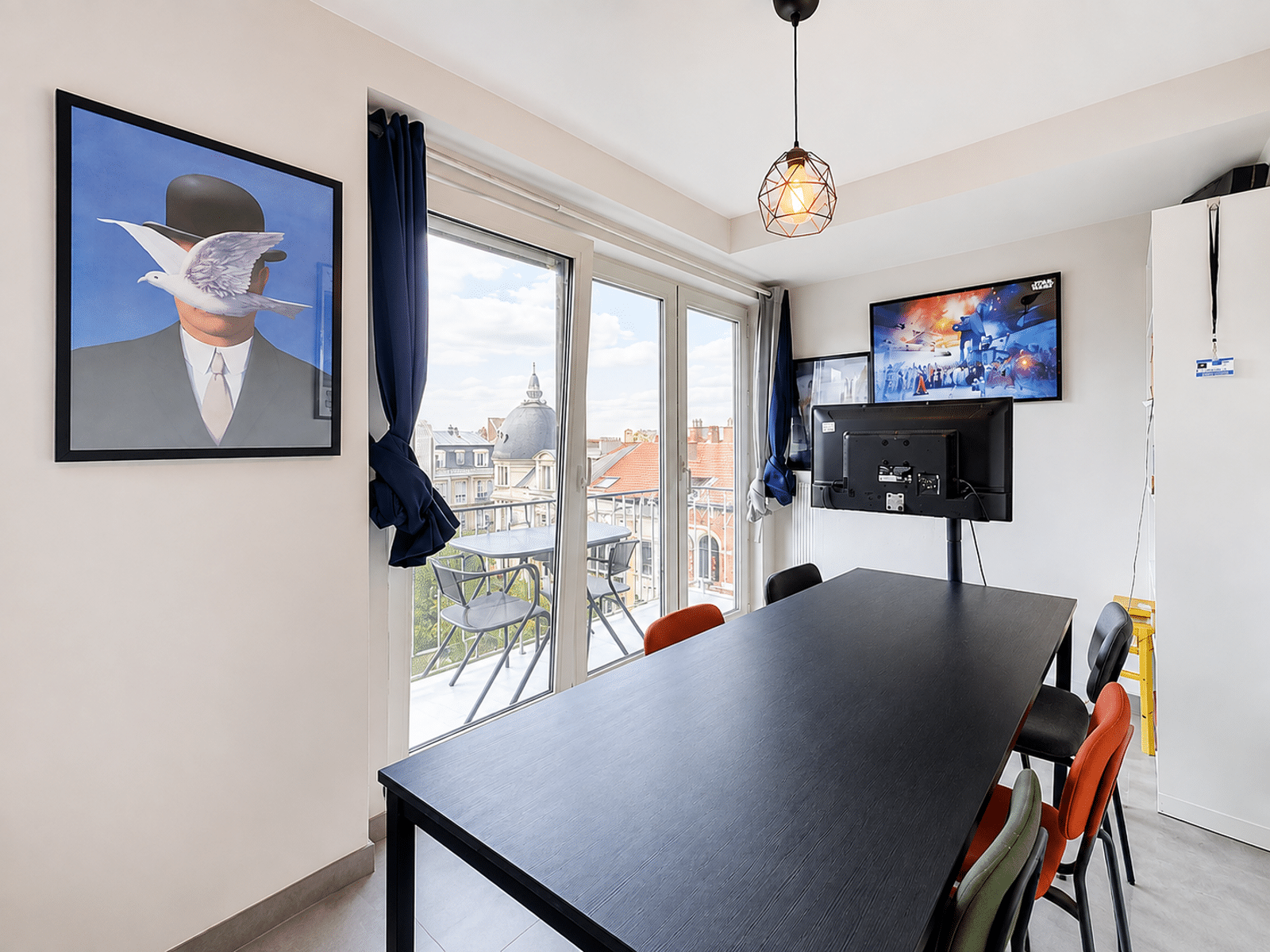 whise photo 4 Conference room with long black table and multicolored chairs, facing a wall-mounted TV showing Star Wars art; large glass doors open to a balcony with city rooftops outside.
