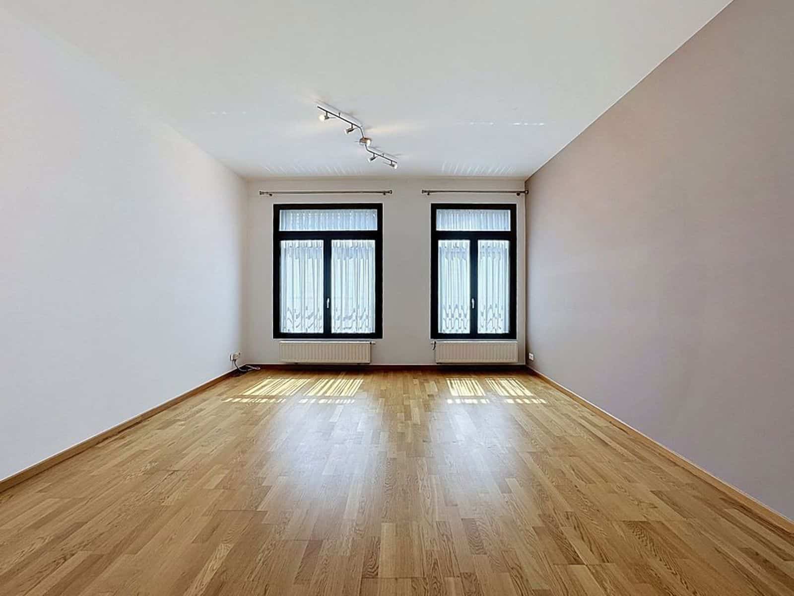Bright living room with parquet floor - You Real Estate Agency A bright, empty living room with hardwood floors, black windows, and radiators in a modern apartment.