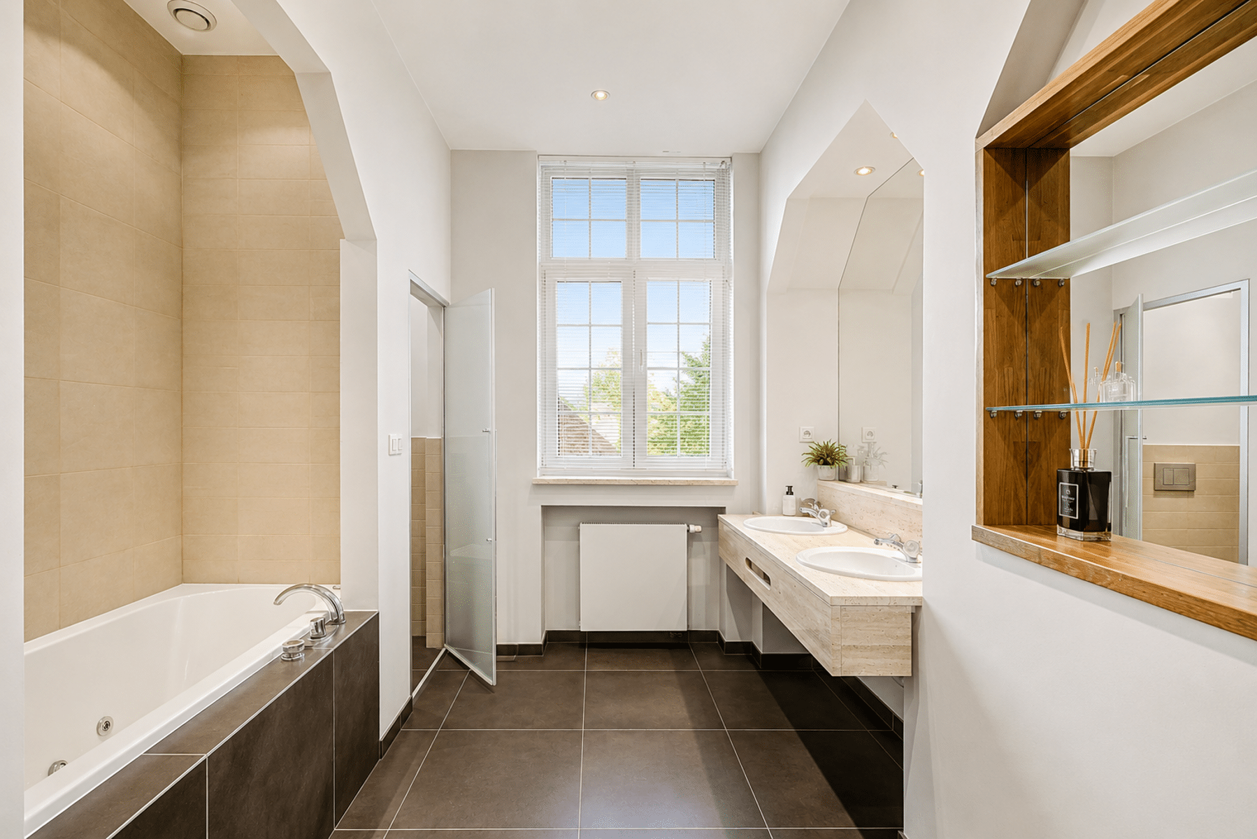 Grote, lichte badkamer – You Real Estate Agency Moderne badkamer met ligbad, dubbele wastafel en groot raam in een licht appartement.