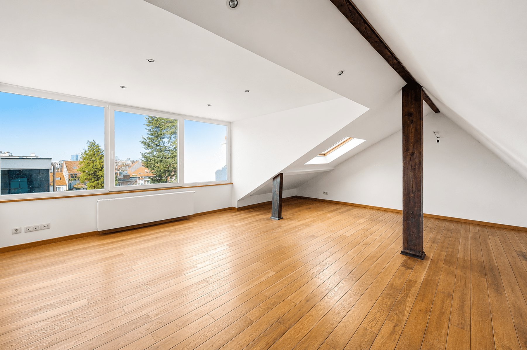 Lichte zolderkamer met balken – You Real Estate Agency Licht zolderappartement met parketvloer, zichtbare balken en grote ramen.