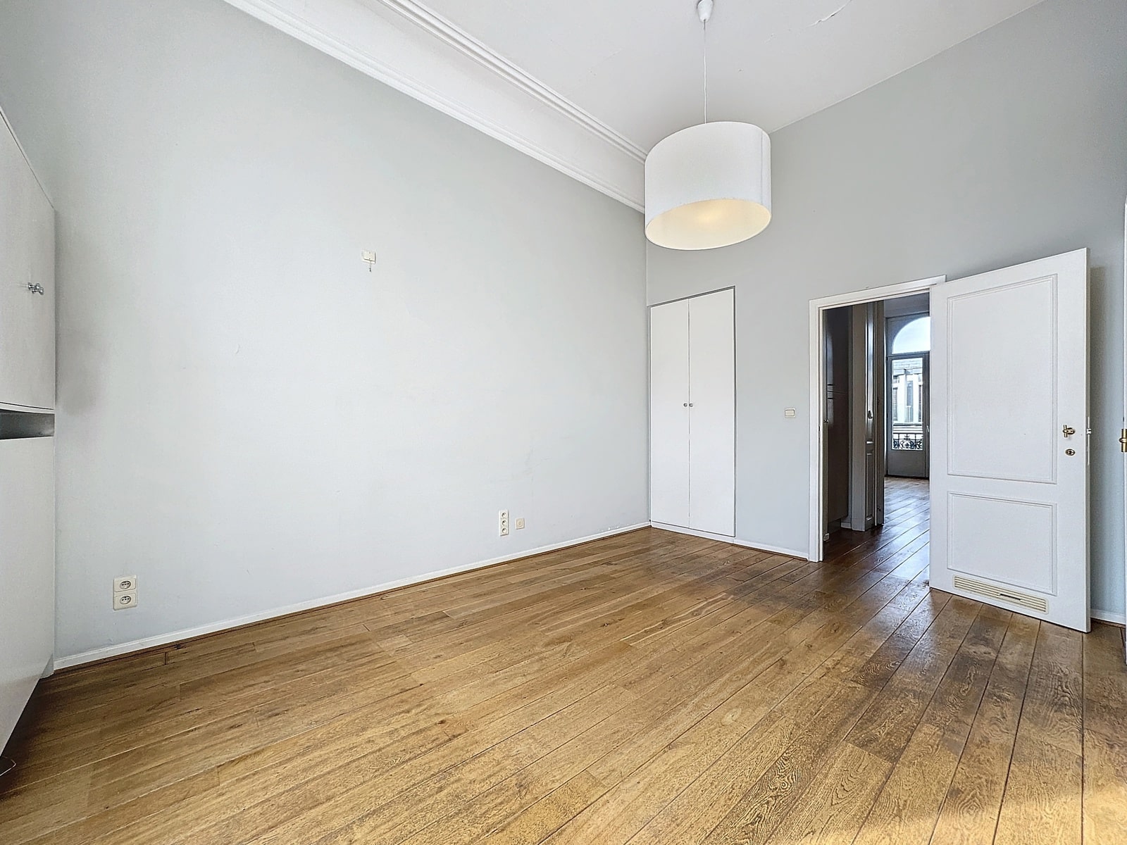 foto 3 Een ongemeubileerde kamer met lichtgrijze muren, witte kroonlijsten en een houten vloer; bij de deuropening hangt een witte, cilindervormige hanglamp aan het plafond.