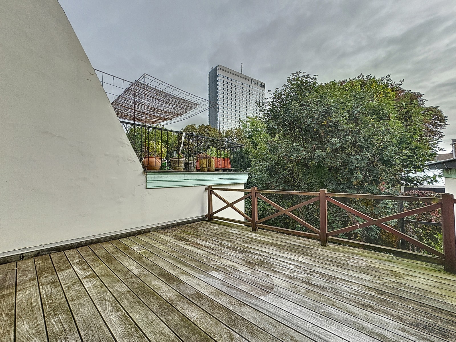 foto 7 Een houten terras met potplanten langs een witte muur, een metalen reling en een grote boom aan de rechterkant; in de verte een hotelgebouw onder een grijze hemel.